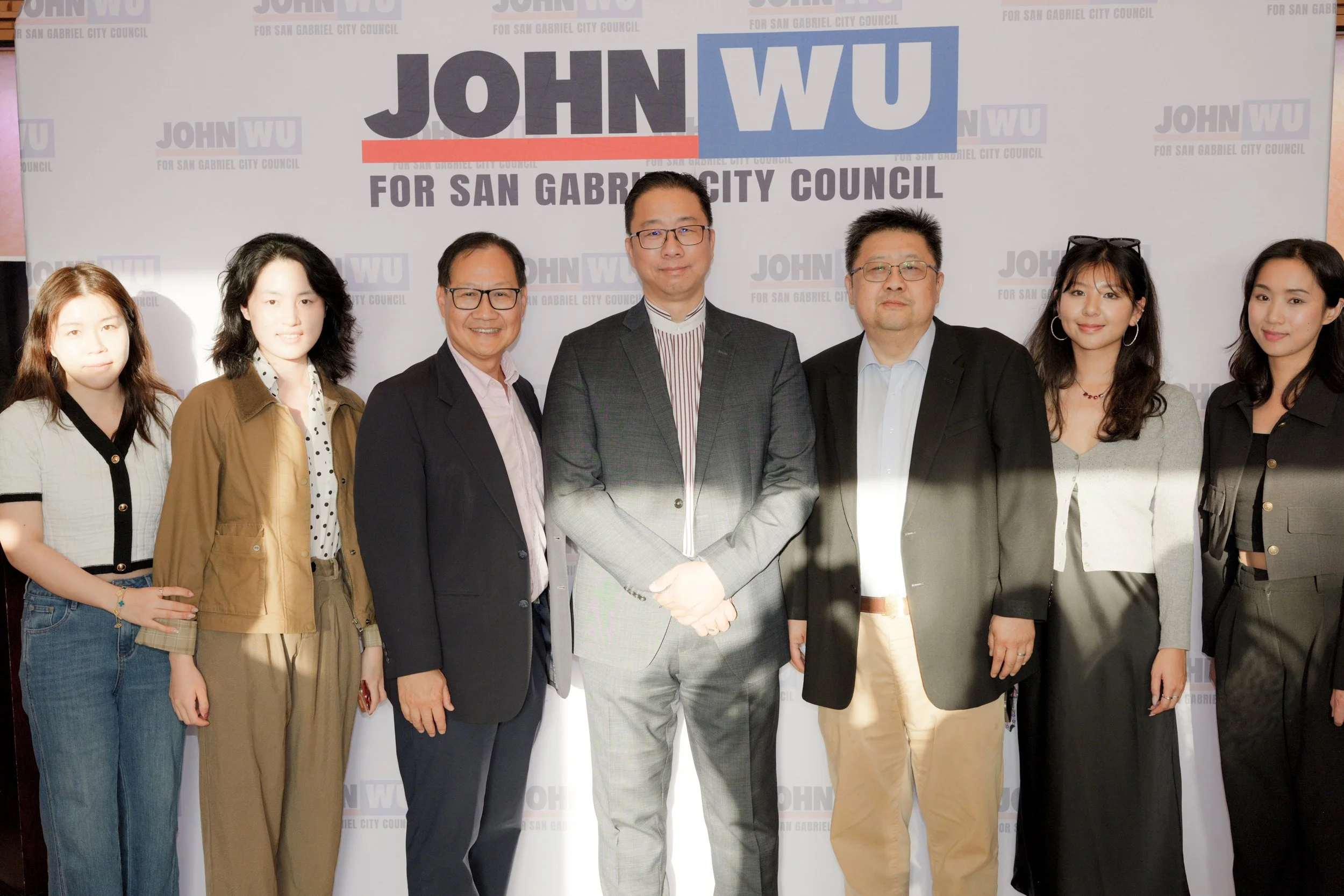 Group of nine diverse people standing together in front of a campaign banner for John Wu running for San Gabriel City Council.