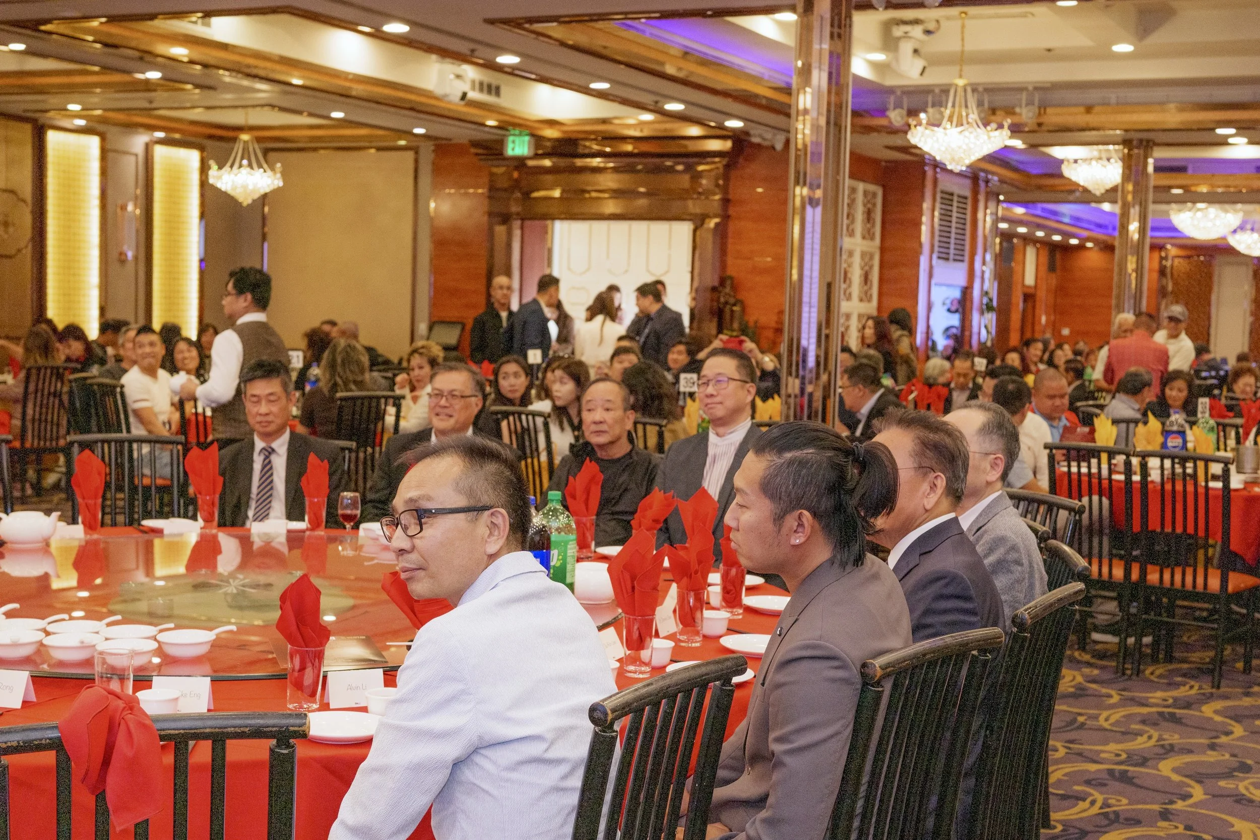A large banquet hall filled with round tables set for a formal event. Many guests are seated, some are talking, and waitstaff are attending to the guests. The room has elegant chandeliers, wooden wall panels, and yellow lighting accents.