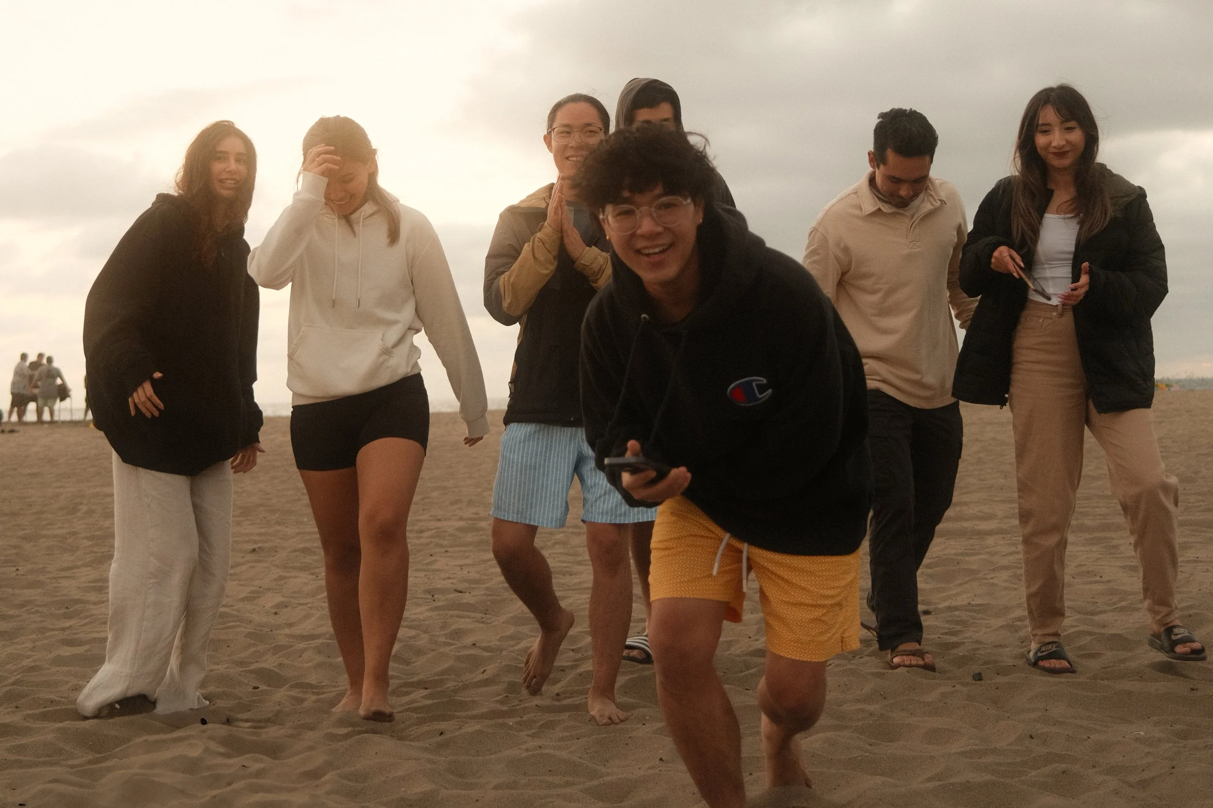 Group of seven young people walking and smiling on a sandy beach on a cloudy day.