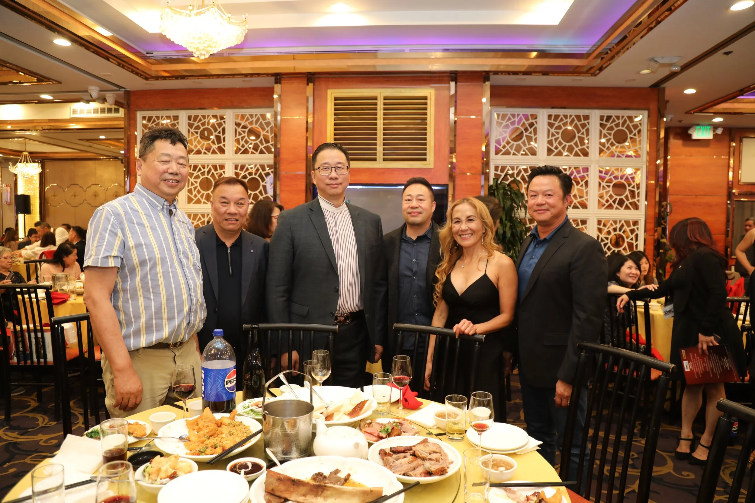 Group of six people, five men and one woman, standing together at a banquet in a restaurant. There is a table with various dishes and drinks in front of them. The restaurant has warm lighting and decorative white lattice panels on the walls.