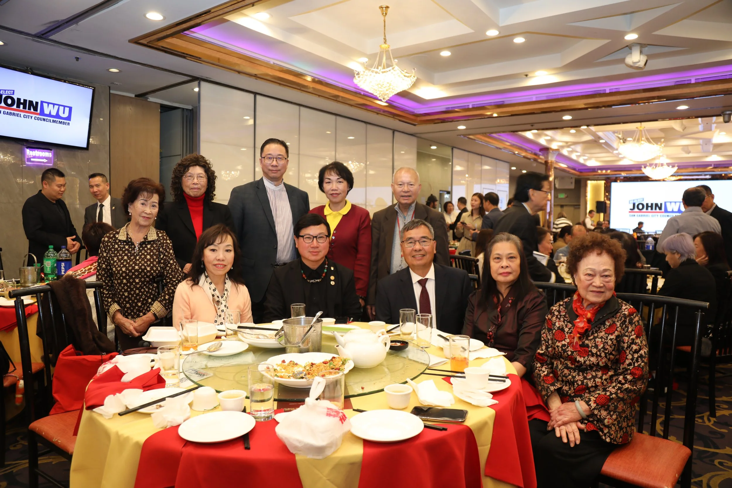 Group of people gathered around a table at a celebration or banquet in a decorated banquet hall with chandeliers and purple accent lighting. Some individuals are standing, others are seated, with plates of food on the table, including leftovers and d