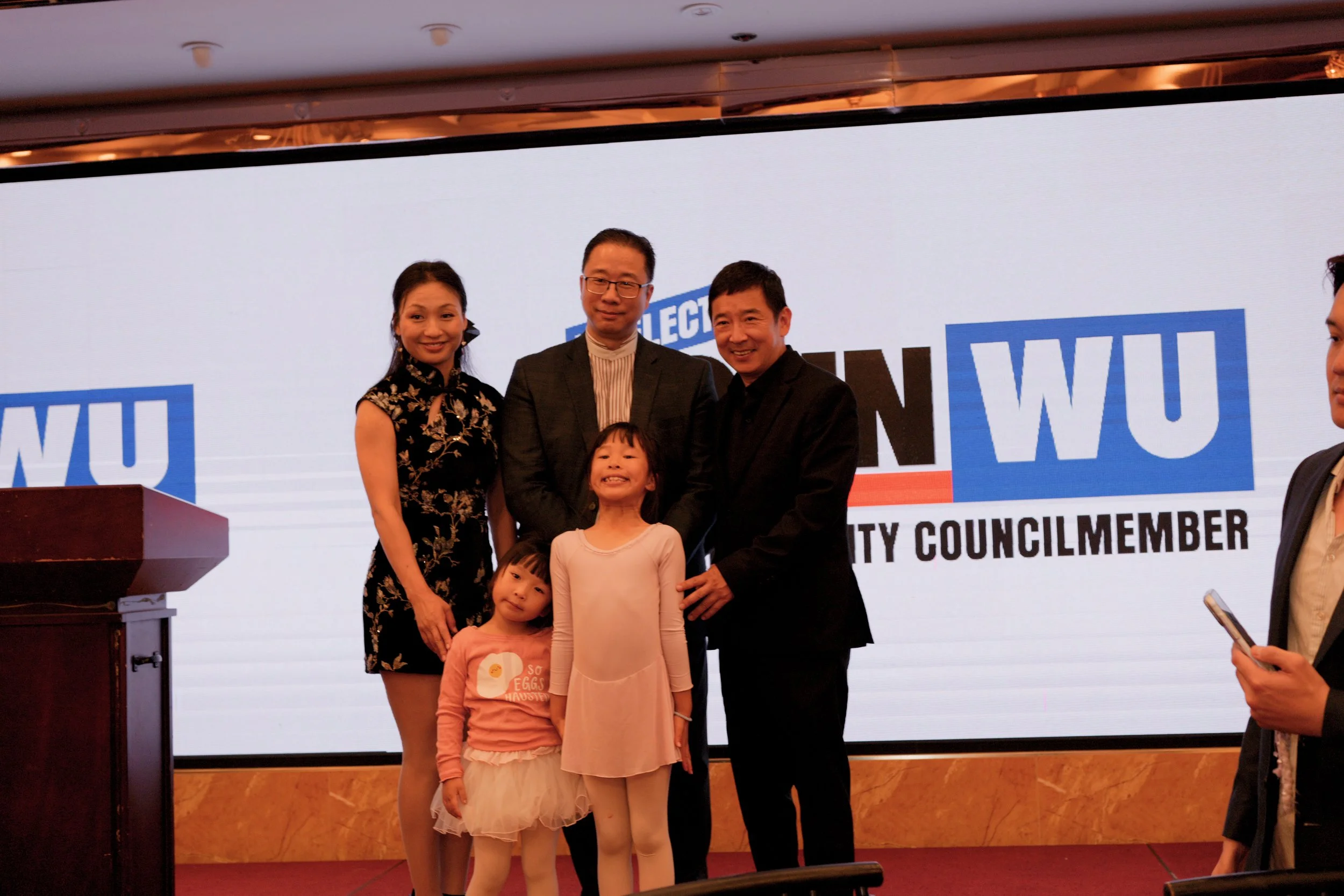 Group of four people, including three children, standing on stage with a large screen behind them displaying the words 'ELECT' and 'COUNCIL MEMBER'.