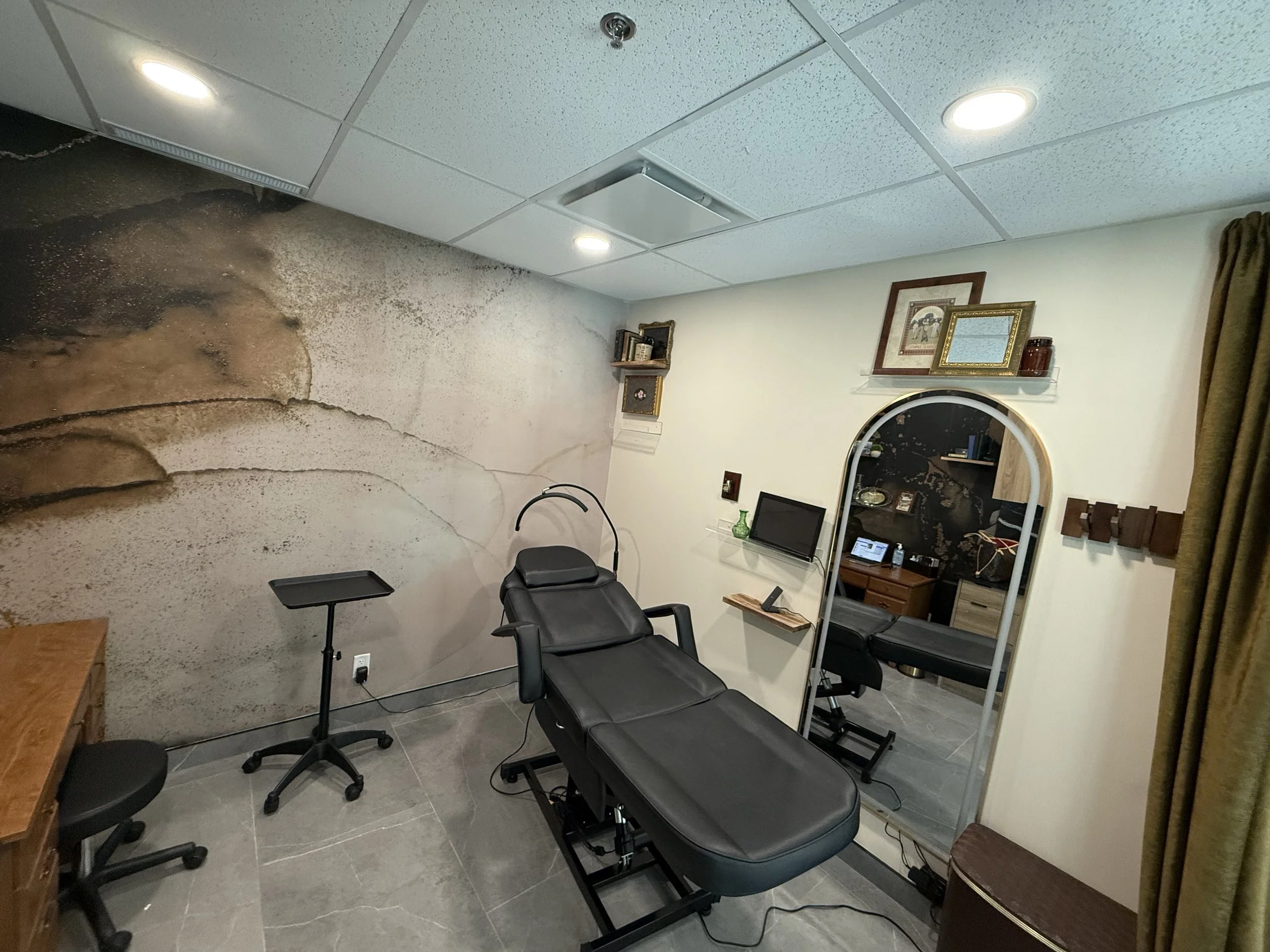 A treatment room with a black examination table, a mirror, a desk with computer, and a decorative wall with a mountain mural.
