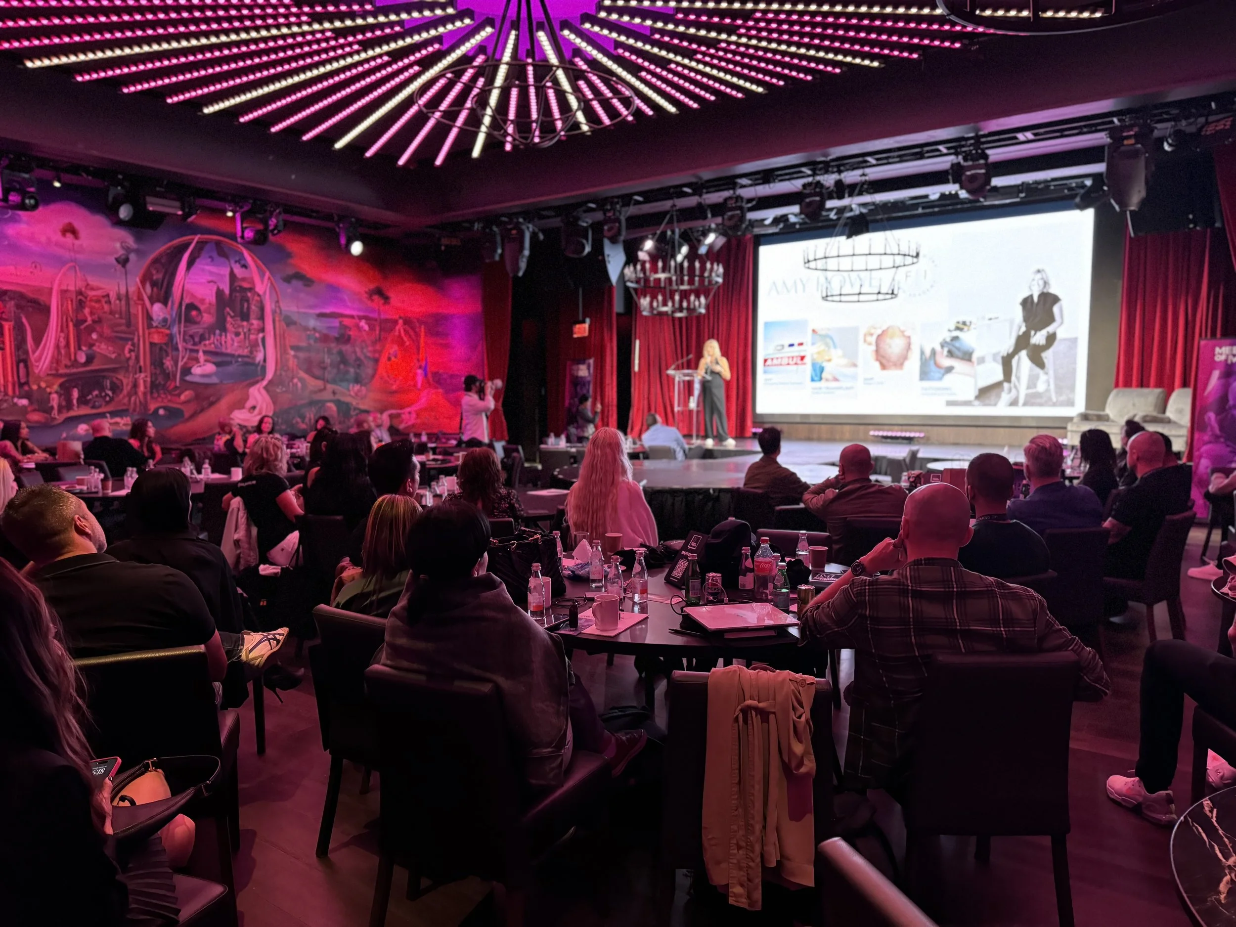 A conference room with an audience watching a woman give a presentation on stage. The room has pink and purple lighting and a large screen displaying slides.
