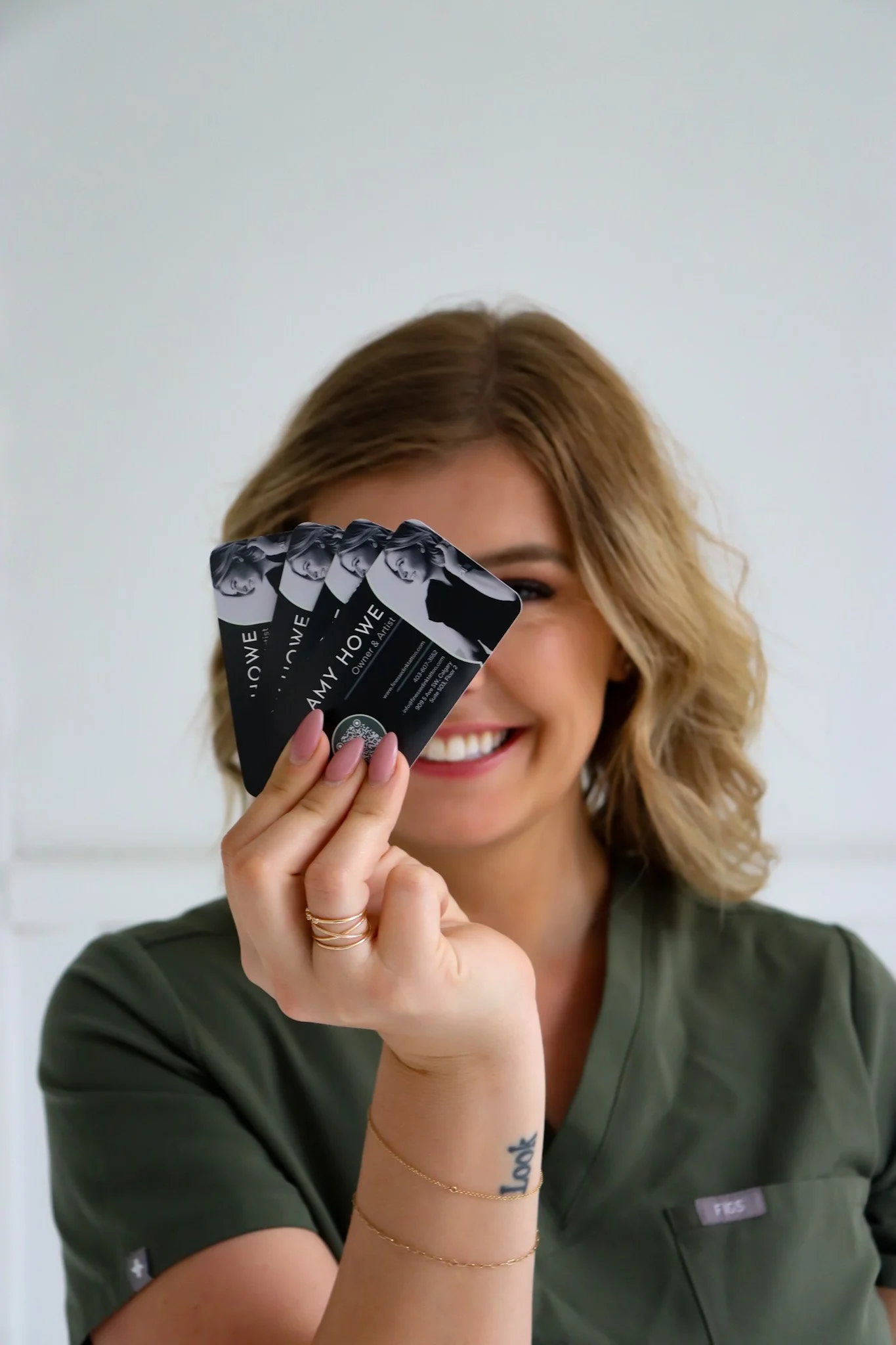 A woman with short wavy hair and a tattoo on her wrist, smiling and holding several business cards with her name 'Amy Howe' in front of her face.