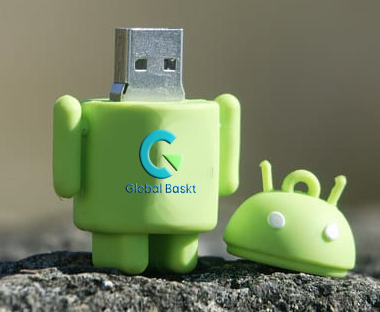 A green Android robot-shaped USB flash drive with the Google logo and the words "Global Bakkt" printed on it, placed outdoors on a rock, with a partially visible Android robot head beside it.