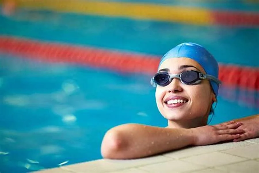 Smiling young girl in swimming cap and goggles resting at the edge of a swimming pool.