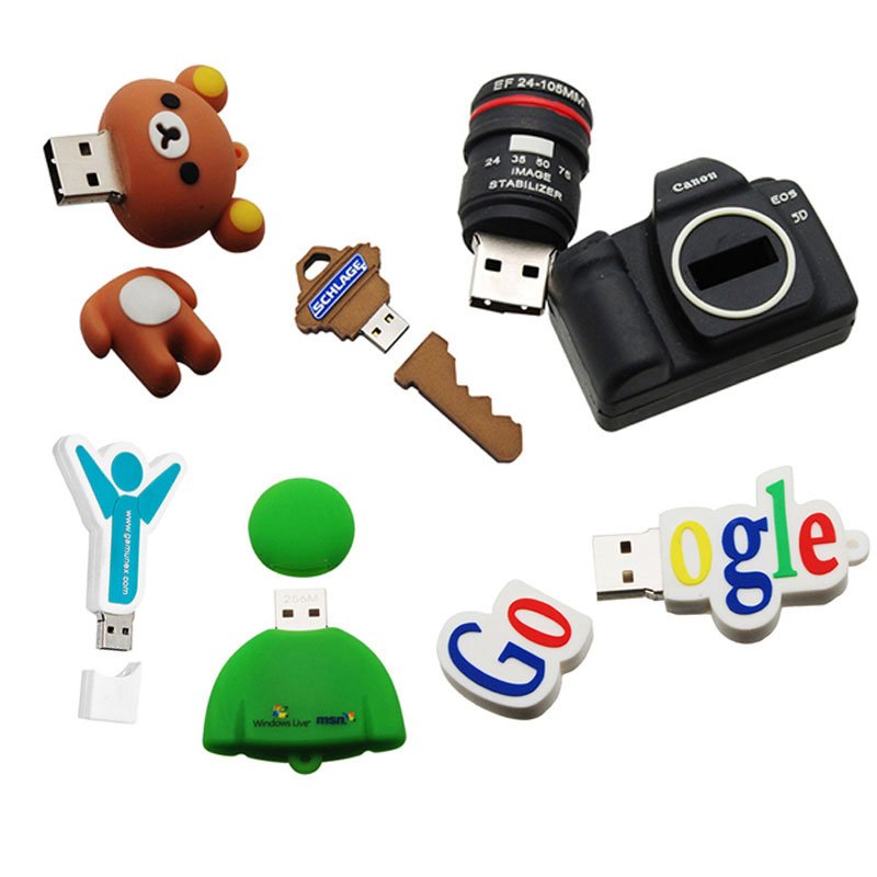 Collection of USB flash drives and camera lens among other electronic accessories and toys, including a bear-shaped USB drive, a brown bear mobile, a gear-shaped USB, a small screwdriver, a camera, a green rubber ball, and Google and Windows logo USB
