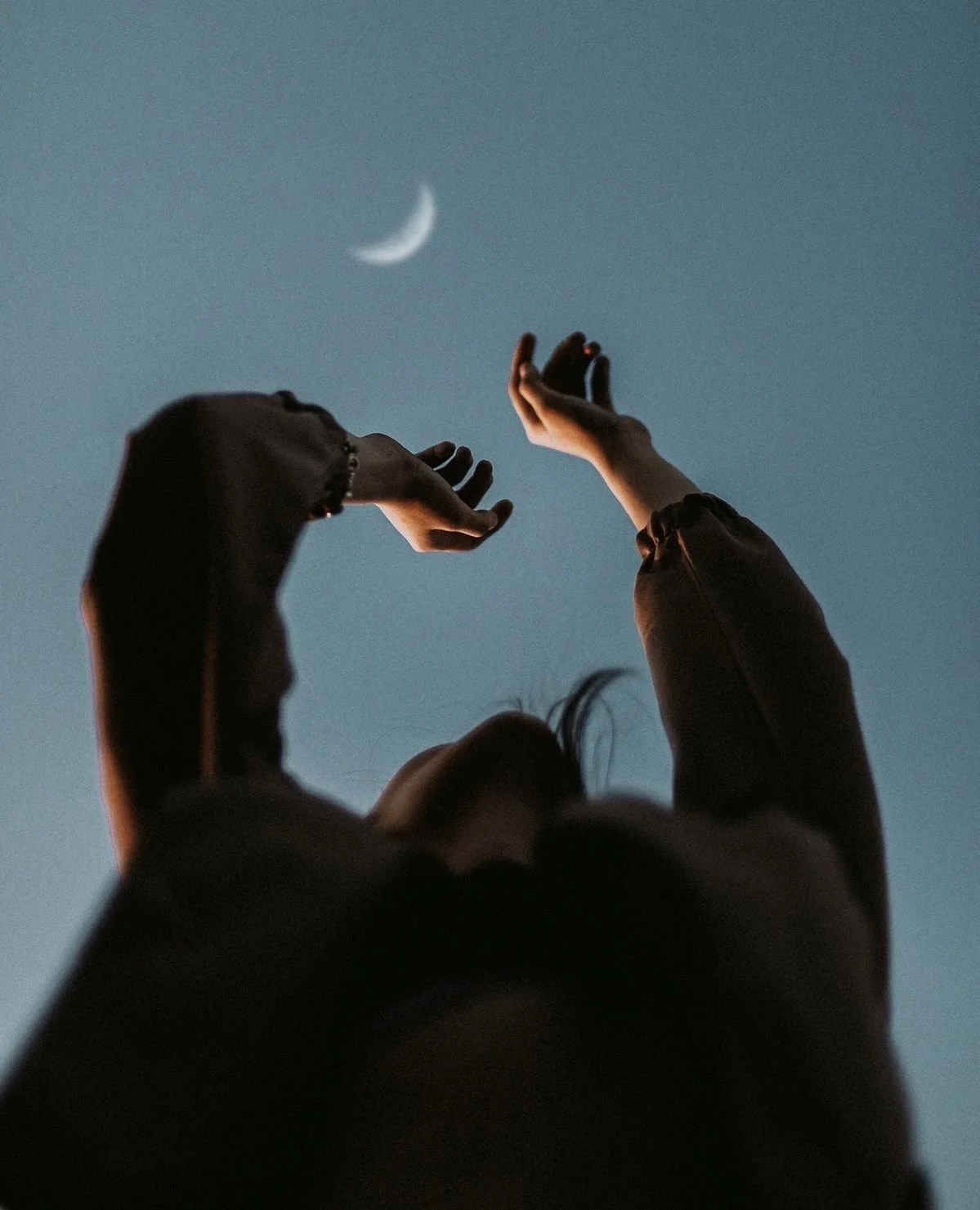 A view looking up at a womans head and chest with her arms raised in the air toward the night sky and crescent moon.