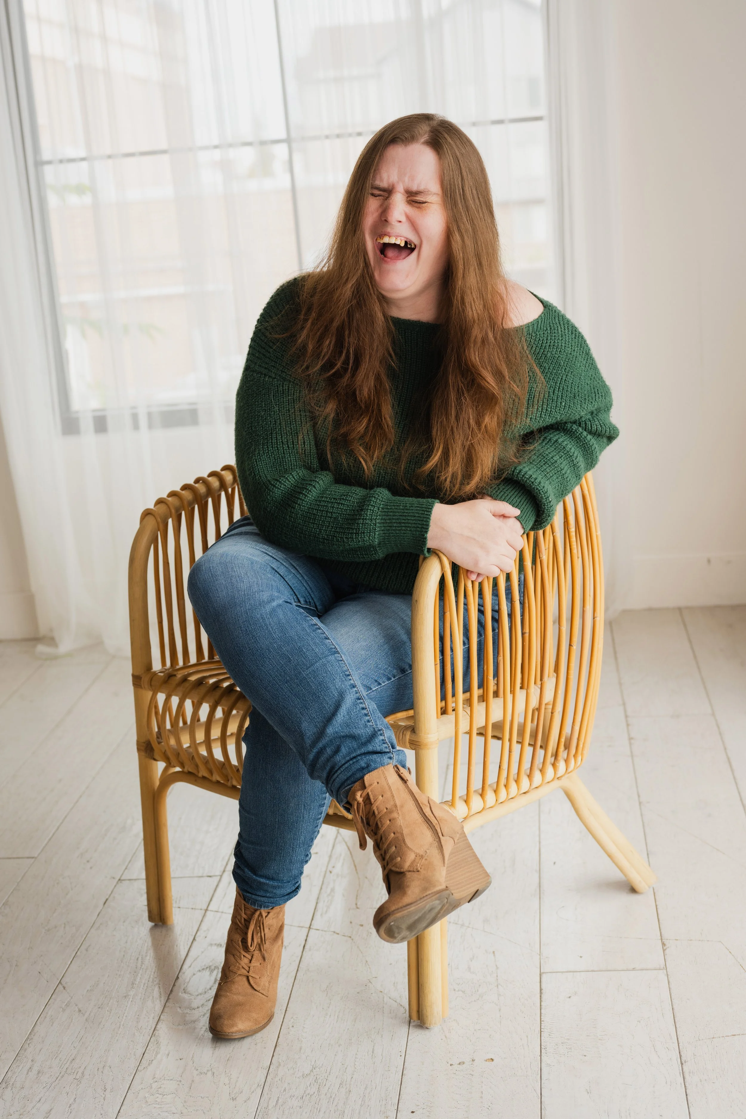 An image of Author Gretchen Elaine, with auburn hair, sitting in a chair laughing and wearing a green sweater.