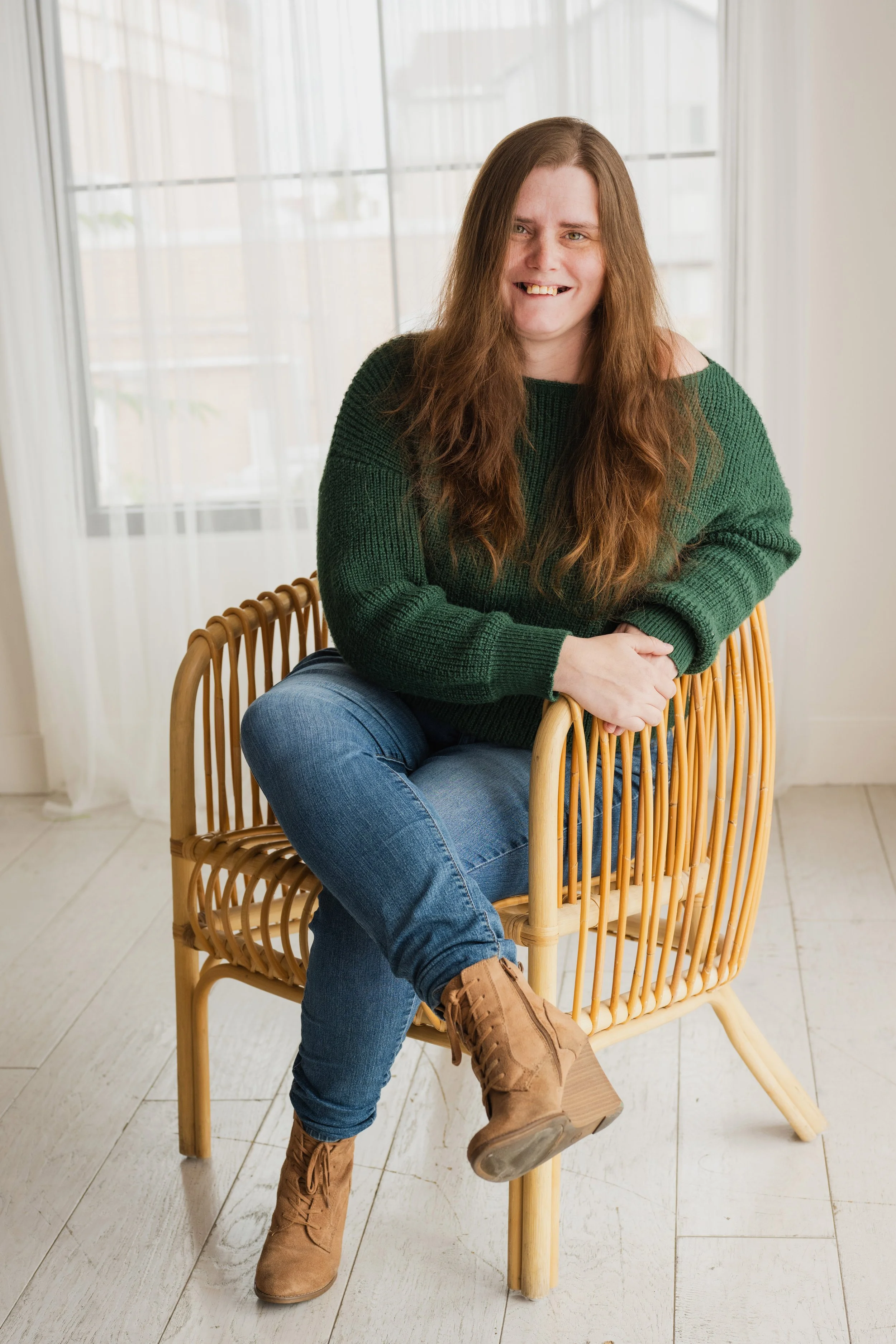 A woman (gretchen elaine) sitting in a rattan chair, wearing jeans and a dark green sweater. She is smiling and her auburn hair drapes in front of her shoulders.