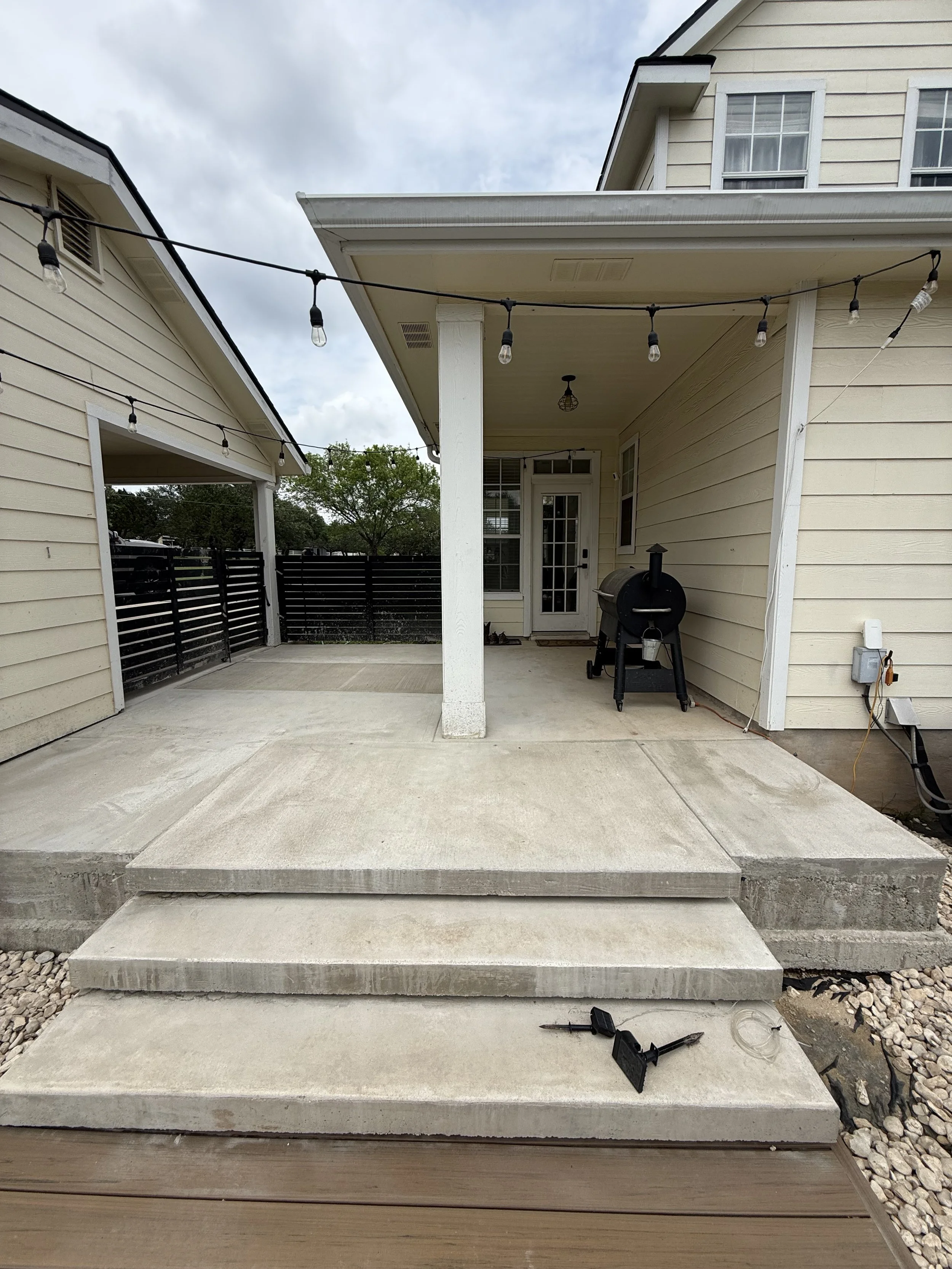 Back porch with three concrete steps leading up to a concrete patio. There are string lights hanging across the porch, a black pellet grill, and a white door with glass panes. The house is light yellow with white trim, and part of a neighboring house is visible on the left.
