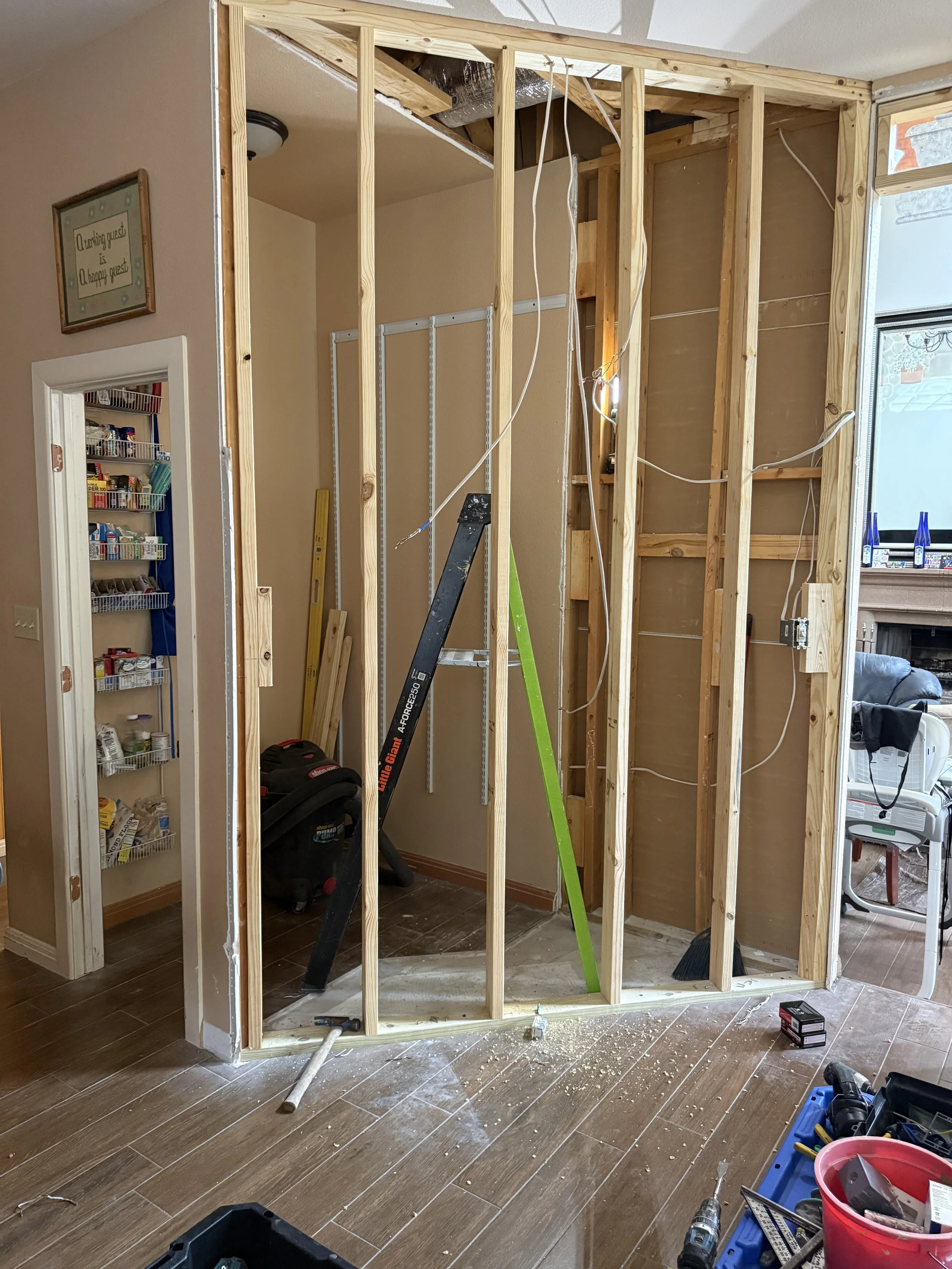 Interior view of a room under renovation with exposed wooden wall studs, construction tools and materials scattered on tile floor, and a small closet or pantry with shelves in the background.