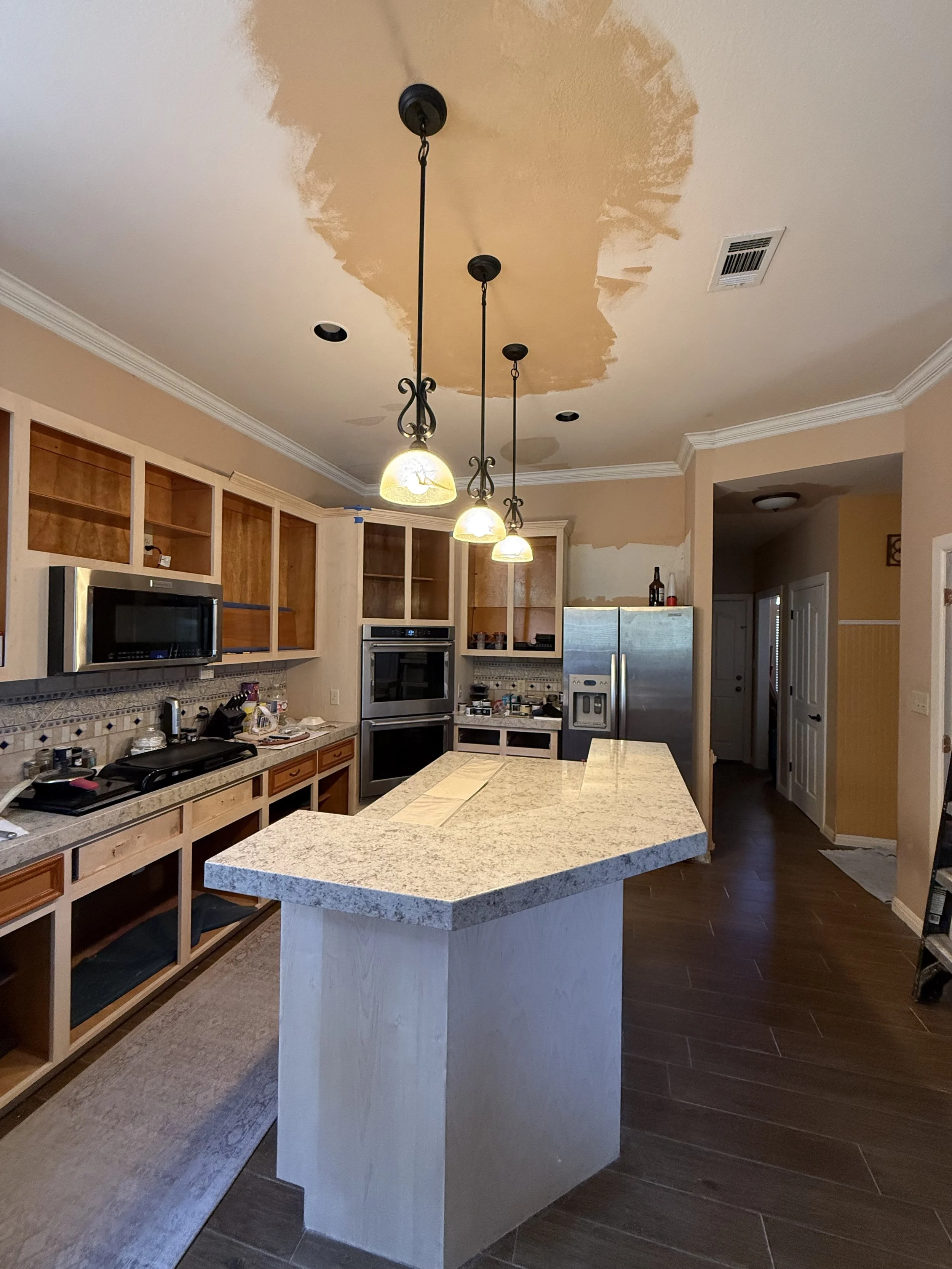 A kitchen with a large island with a granite countertop, wooden cabinets, and built-in appliances. The ceiling has an unfinished patch of paint or drywall, with three hanging pendant lights.