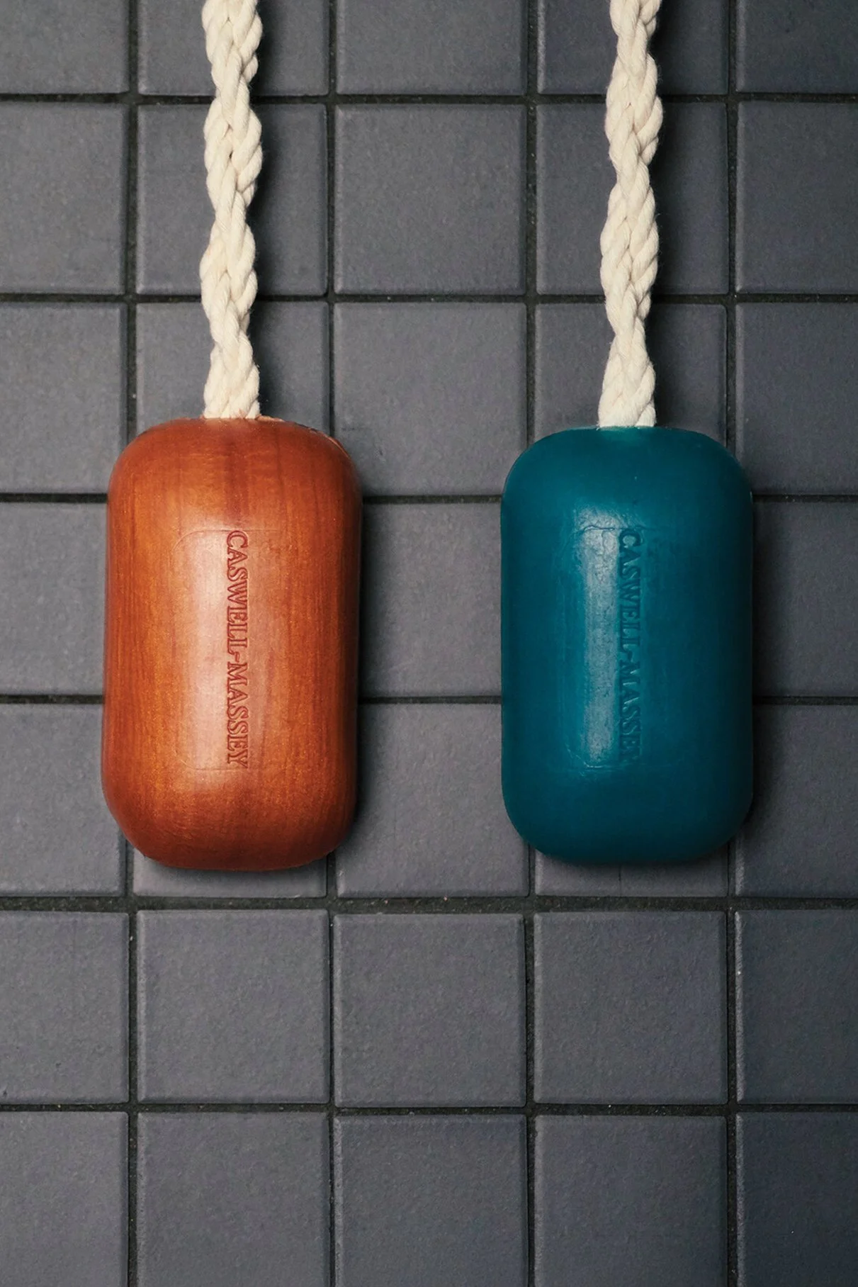 Two massage tools, one brown and one blue, hanging on a gray tiled wall, with white rope handles.