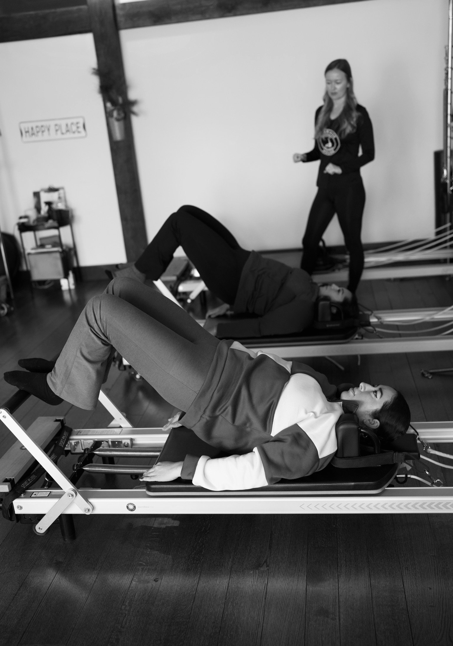 Two women exercising on Pilates reformer machines in a studio with an instructor watching.