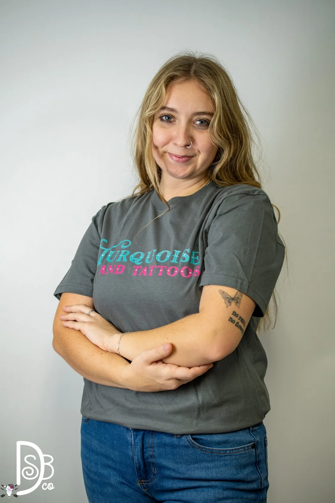 A woman with blonde hair, a nose ring, and tattoos, wearing a gray T-shirt with the words "TURQUOISE AND TATTOOS," standing against a plain background.