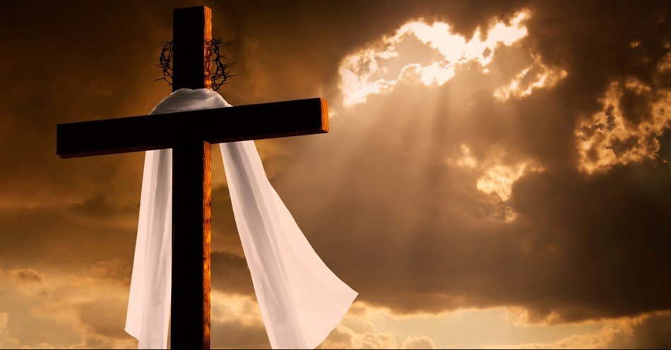 Cross with white cloth or ribbon draped on it, against a dramatic sunset sky with clouds and sunlight shining through.