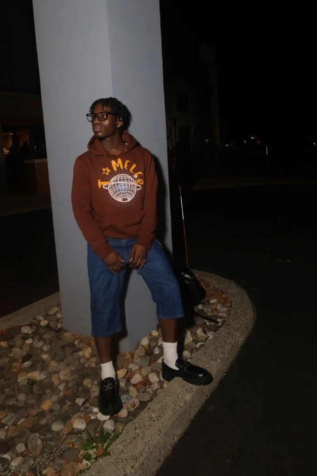 A young person standing outside at night, leaning against a white pillar. They are wearing glasses, a brown hoodie with a globe and star design, baggy jeans, white socks, and black shoes. There is a black bag on the ground next to them.