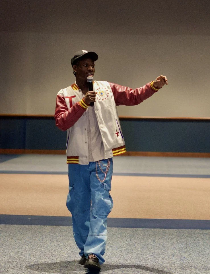 A person standing on a stage, holding a microphone and speaking to an audience. They are wearing a letterman jacket, a cap, and baggy jeans.