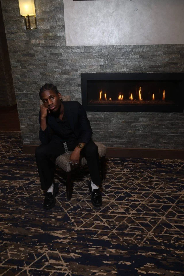 Person sitting on a chair in front of a modern fireplace with flames, against a stone wall in an indoor setting.