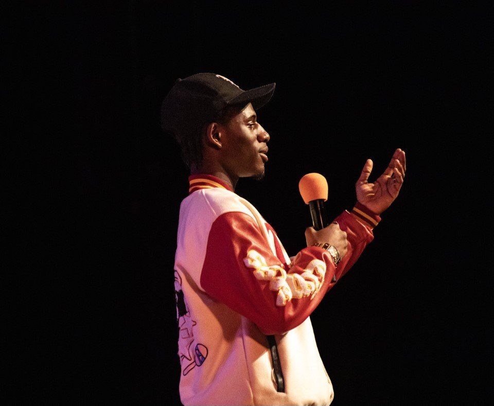 A young man standing on stage, speaking into a microphone with an orange foam cover, wearing a black baseball cap and a red and white jacket, with a dark background.