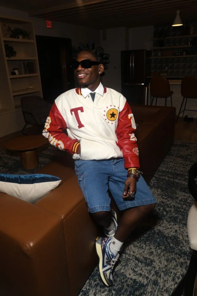 Young man sitting on a brown leather couch with one leg crossed over the other, wearing sunglasses, a colorful varsity jacket, denim shorts, sneakers, and accessories, in a modern living room.
