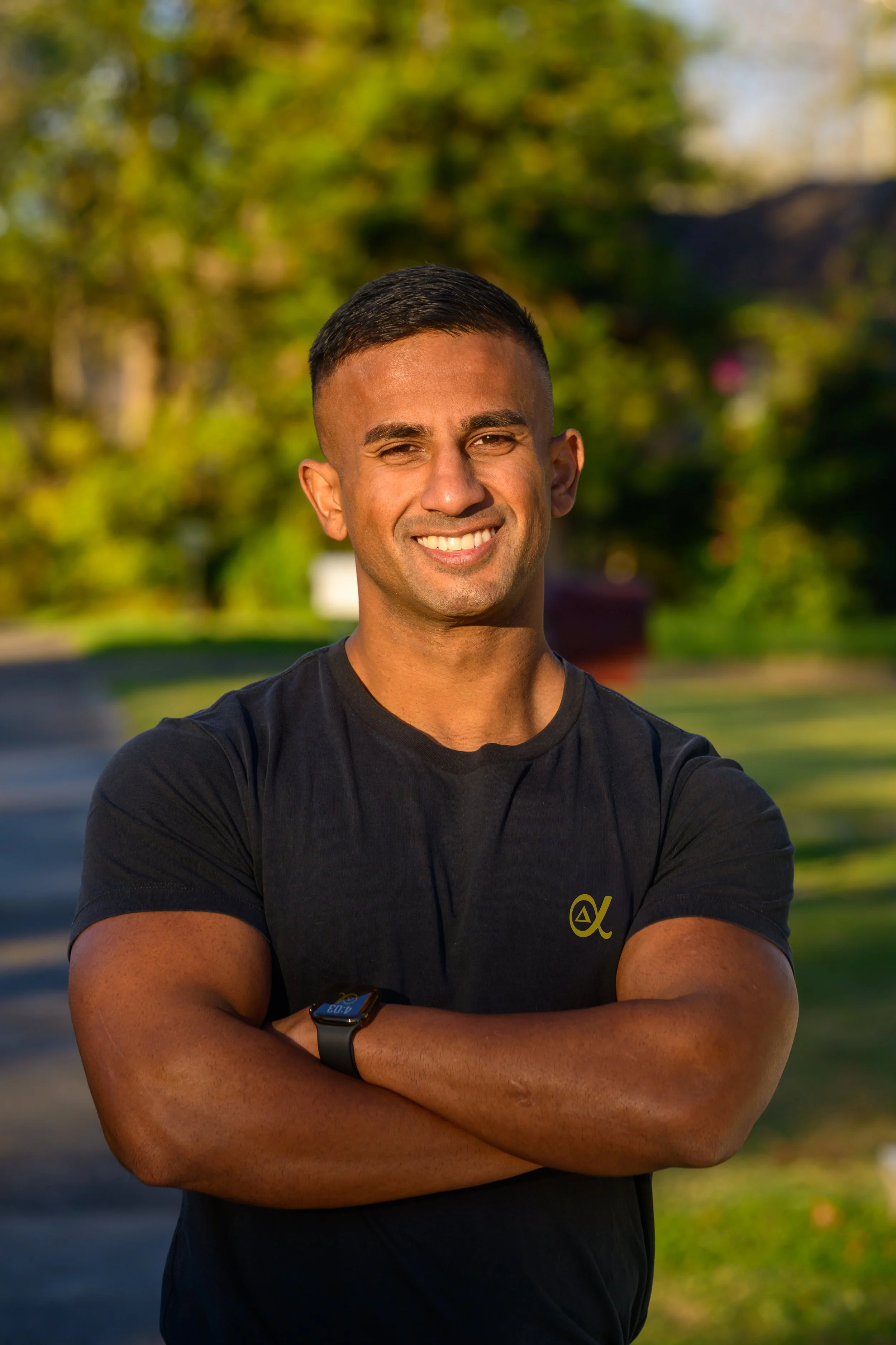 Smiling man with crossed arms, wearing a black t-shirt and smartwatch, outdoors with green trees in the background.