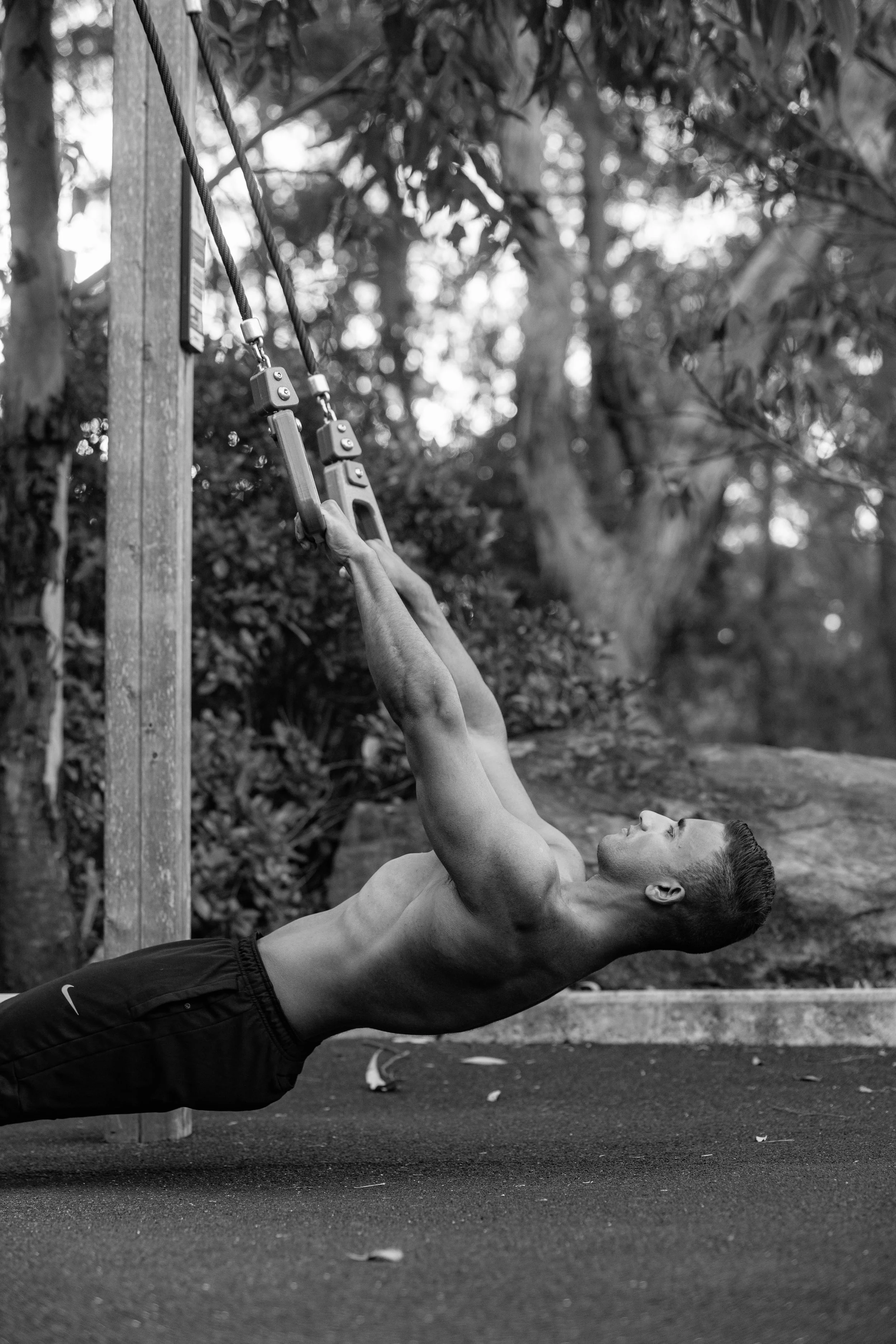 A shirtless man performs a row exercise using suspension straps outdoors, with trees and natural scenery in the background.
