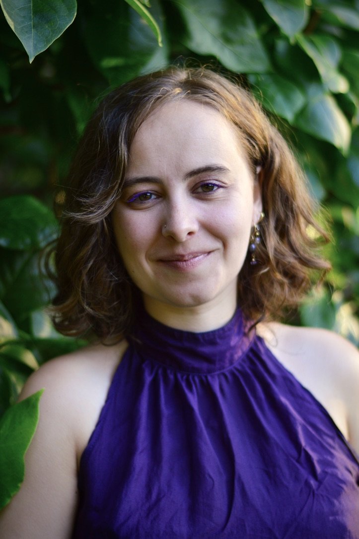 A woman with shoulder-length wavy brown hair, wearing a purple sleeveless top, smiling softly, with green leaves in the background.