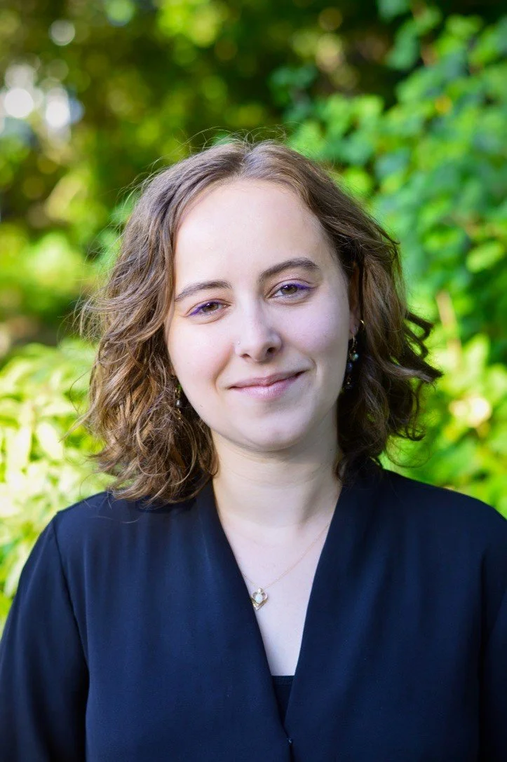 A person with shoulder-length curly brown hair smiling outdoors with green foliage in the background.