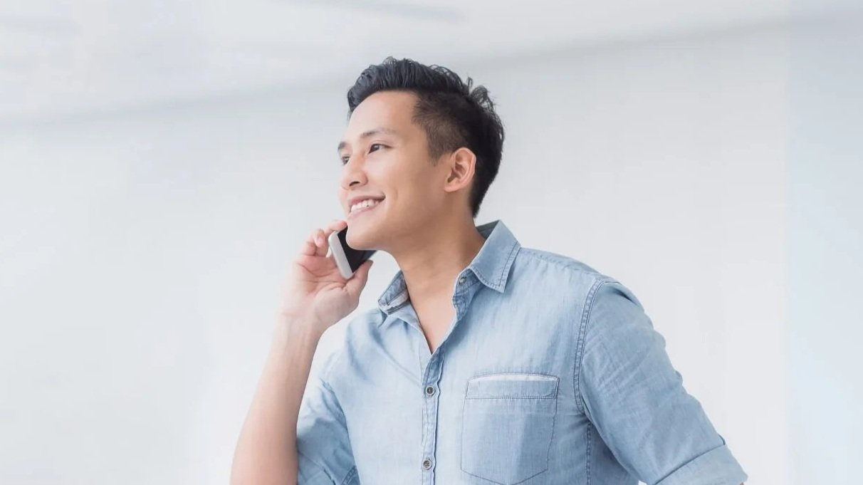 Young man smiling and talking on a cellphone, wearing a light denim shirt, standing against a plain background.