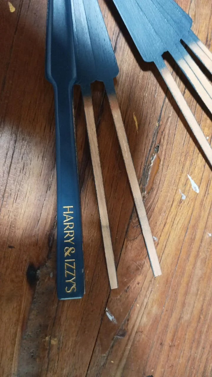 Close-up of a blue spatula with gold lettering that reads "HARRY & IZzy's" and some wooden utensils on a wooden surface.