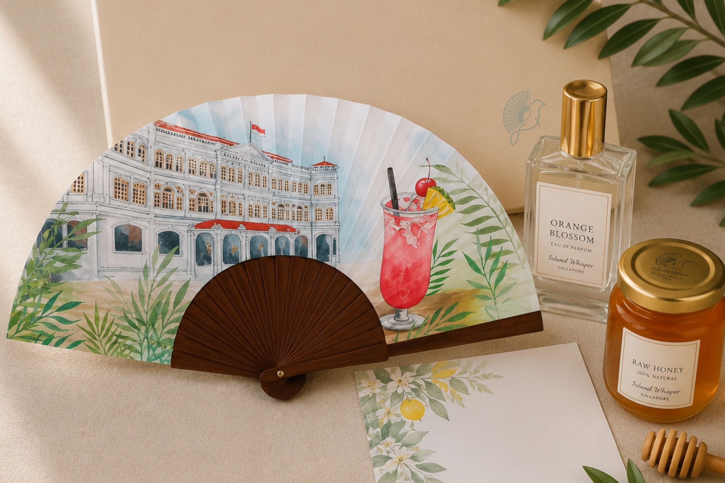 Abanico decorativo con ilustración de un edificio colonial y una bebida con cerezas y rodajas de limón, acompañado de frascos de perfume y miel encima de una mesa beige con hojas verdes y un ramo de plantas.
