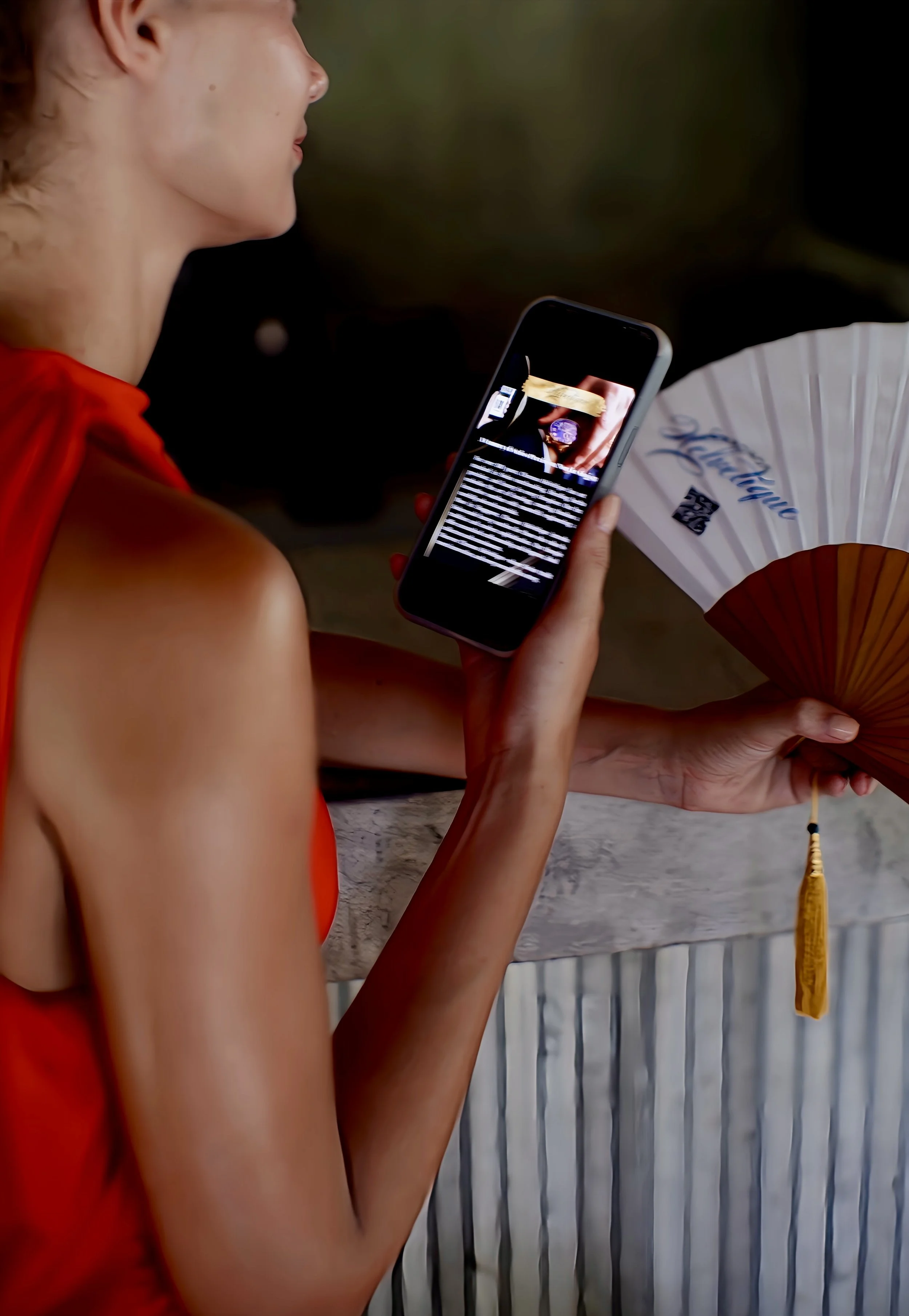 A person in a red dress holding a smartphone in one hand and a decorative fan with a tassel in the other hand, standing by a textured surface.