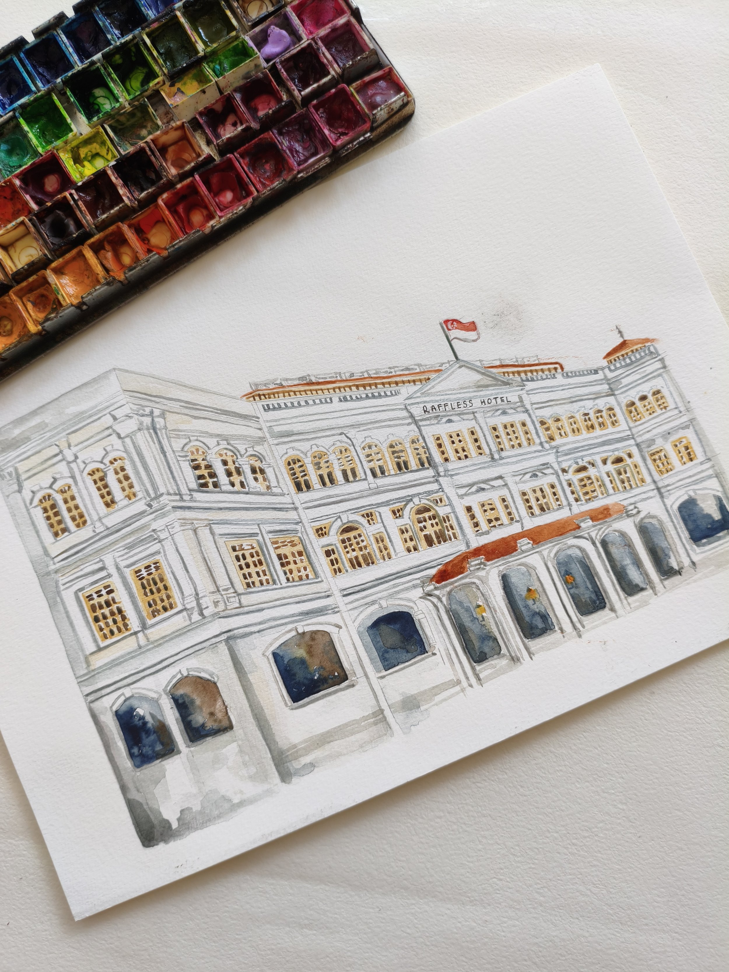 Dibujo colorido de un edificio con ventanas y una bandera, acompañada de una paleta de acuarelas con diferentes colores.