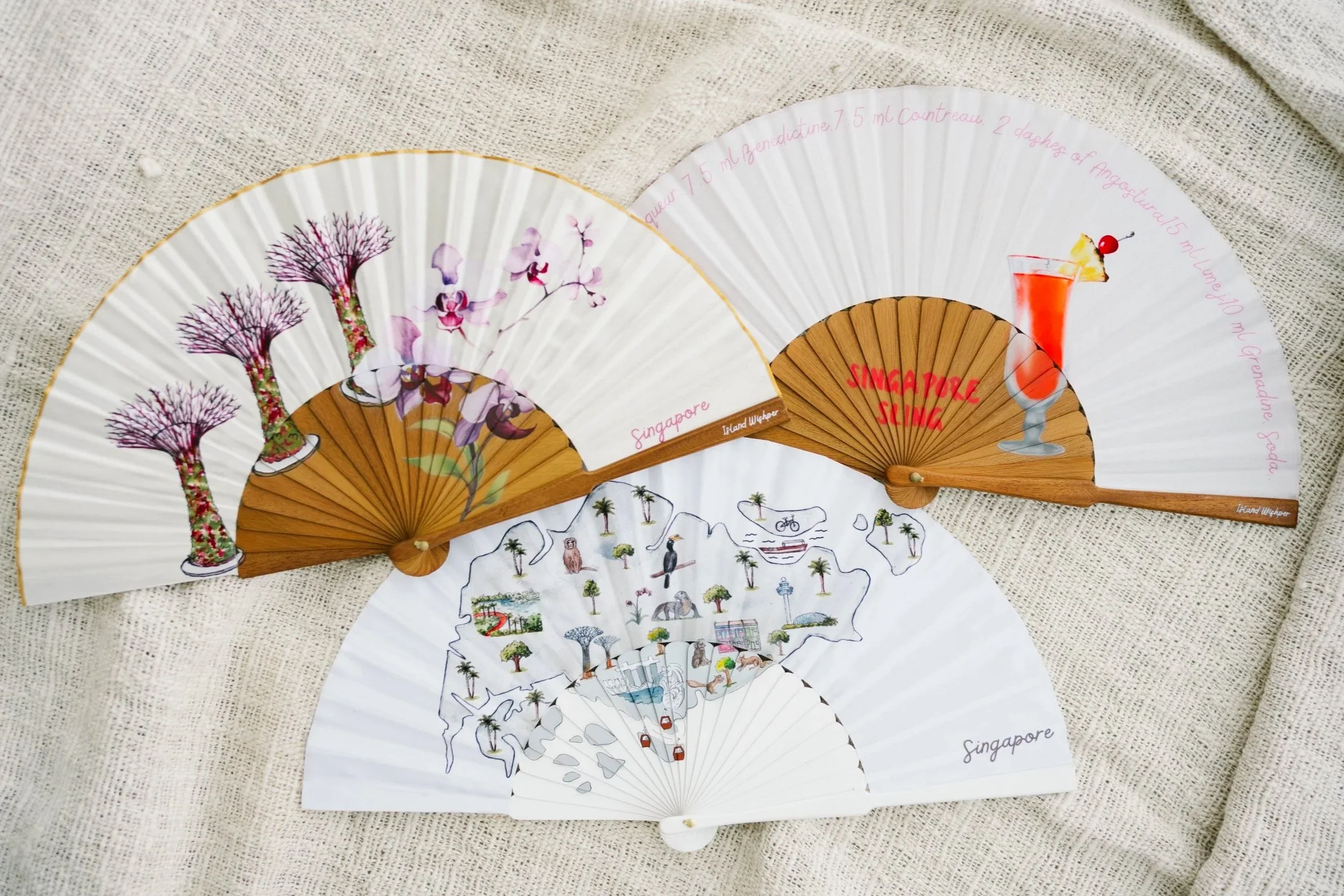 Three Island Whisper Atelier hand fans inspired by Singapore, featuring Gardens by the Bay, orchids, the Singapore Sling cocktail and an illustrated city map.