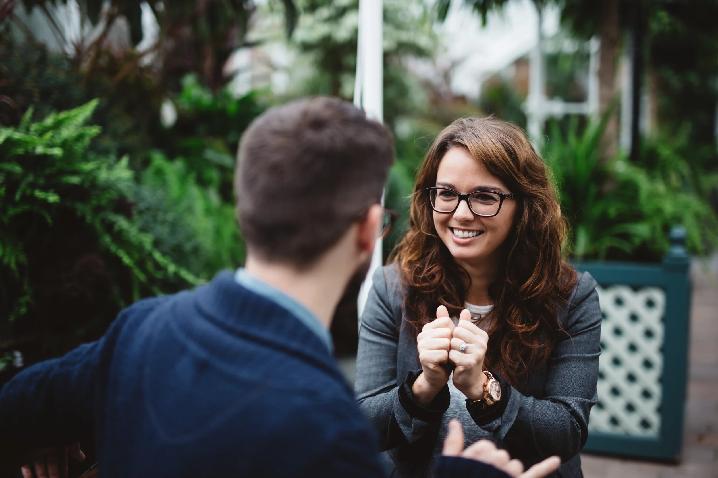 A woman with glasses and long wavy hair smiling and using sign language to say together with a man whose back is to the camera, in a lush outdoor garden setting.