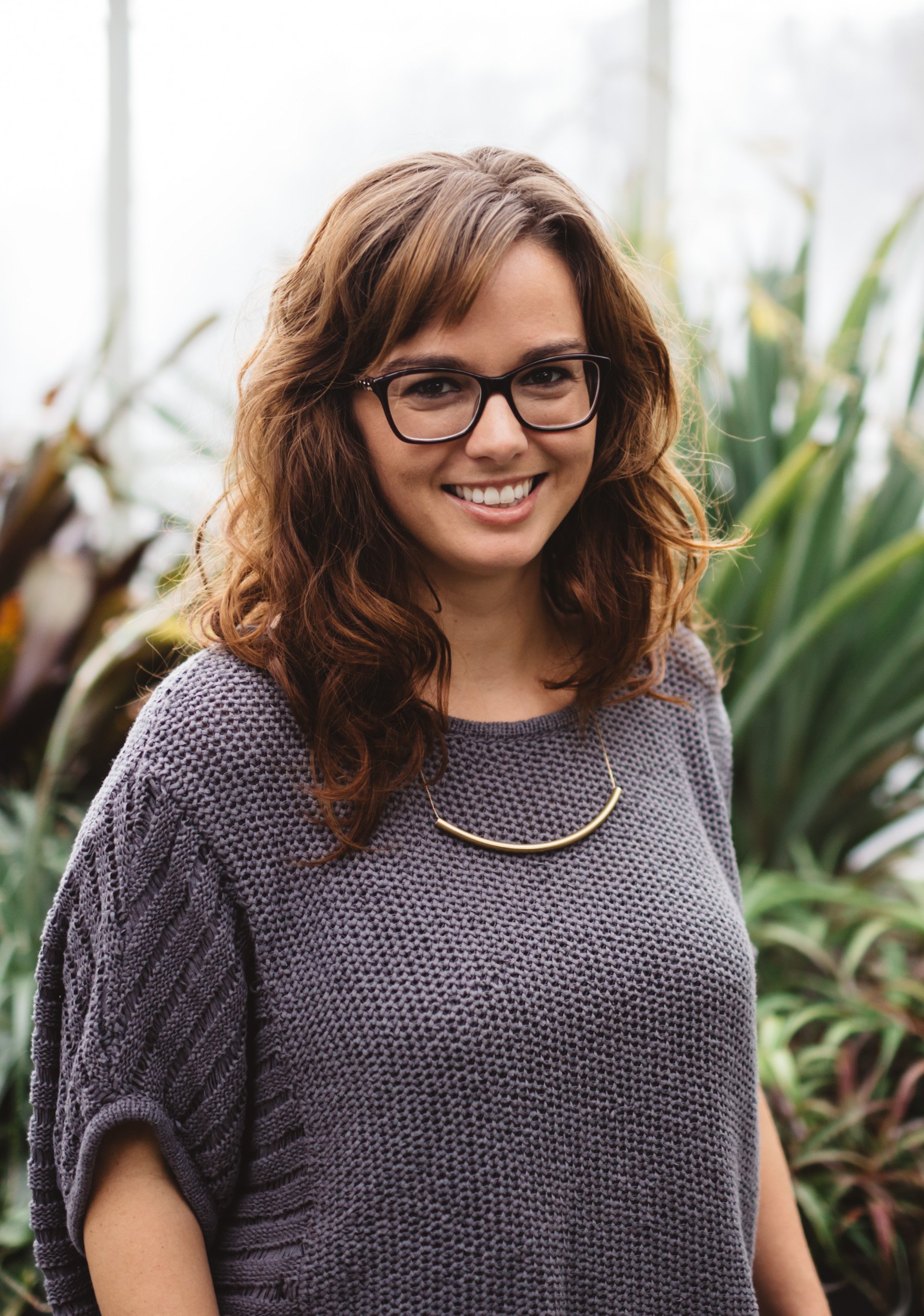 A woman with brown hair, glasses, and a gray knit sweater smiling indoors with green plants in the background.