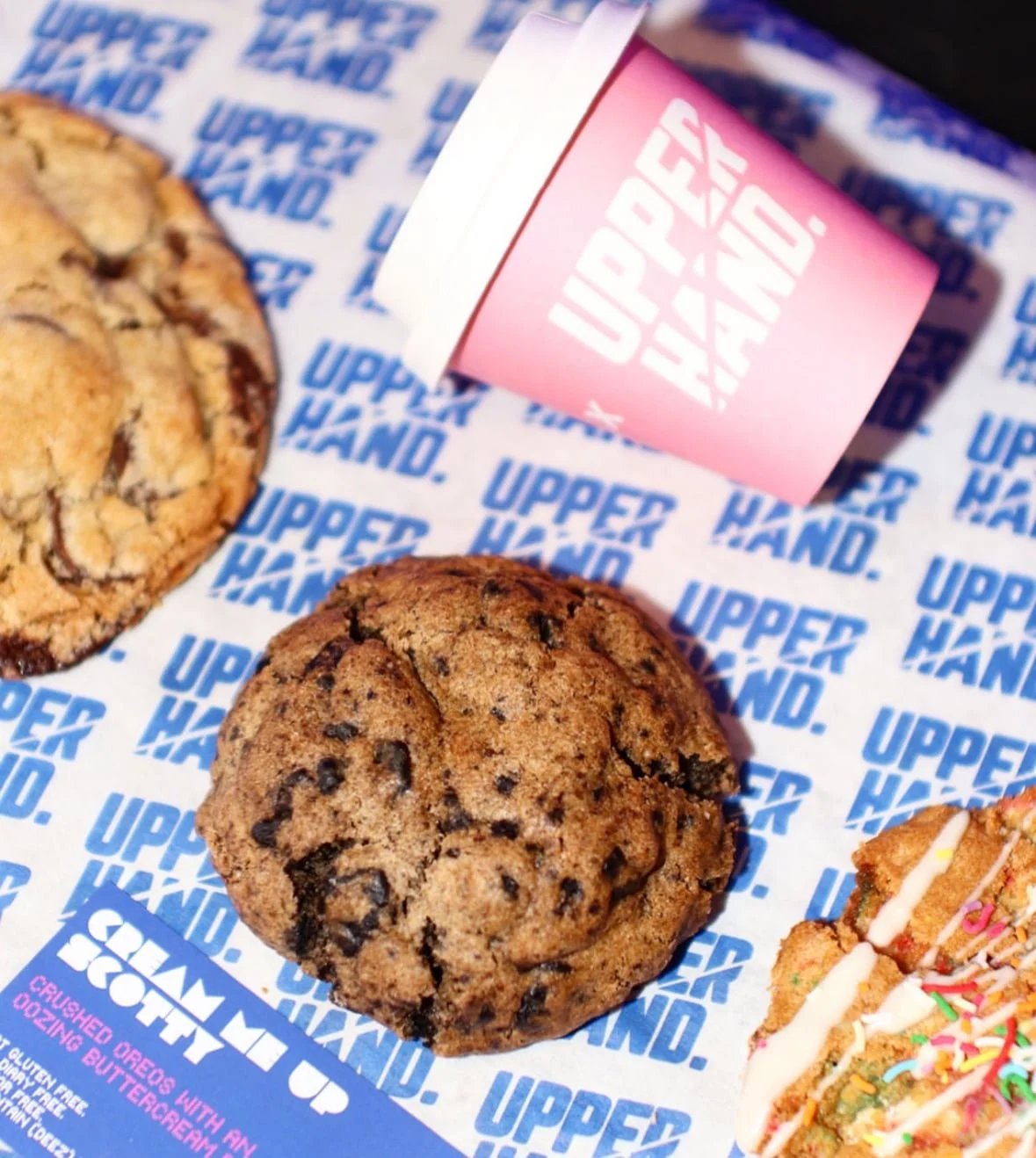 Several cookies, including a chocolate chip cookie, in a box from Upper Hand. There is also a pink cube with white text, and a portion of a sugar cookie with colorful sprinkles.