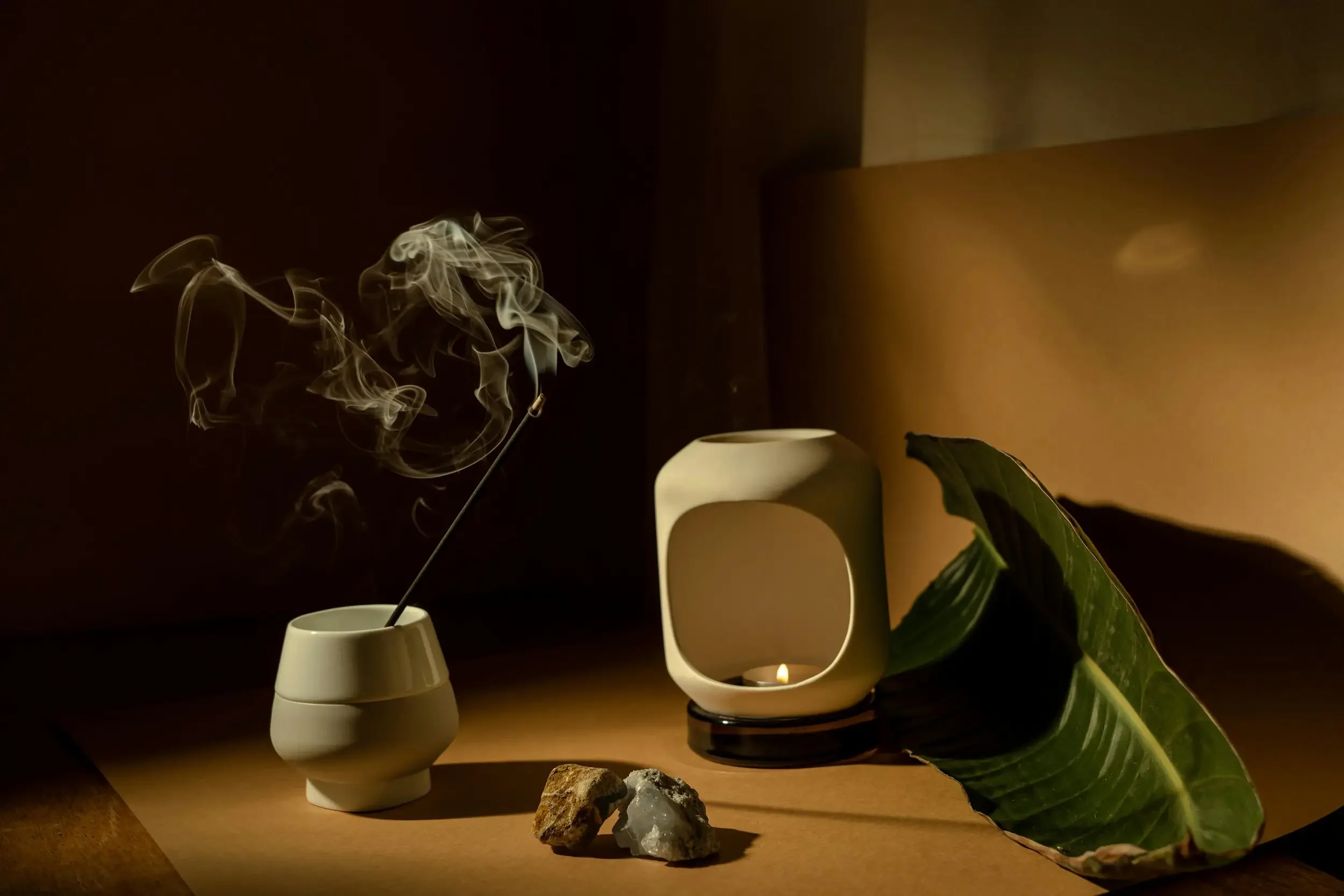 Stylish interior scene with incense burner emitting smoke, white ceramic candle holder, rocks, and a large green leaf on a brown background.