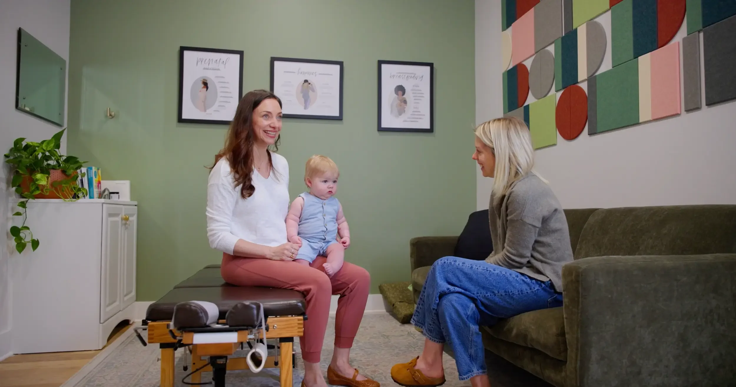 A woman holding a baby sits on a chiropractic table in a room with green walls, while another woman sits on a sofa. The room has framed posters and decorative wall art. A shelf with books and plants is visible.