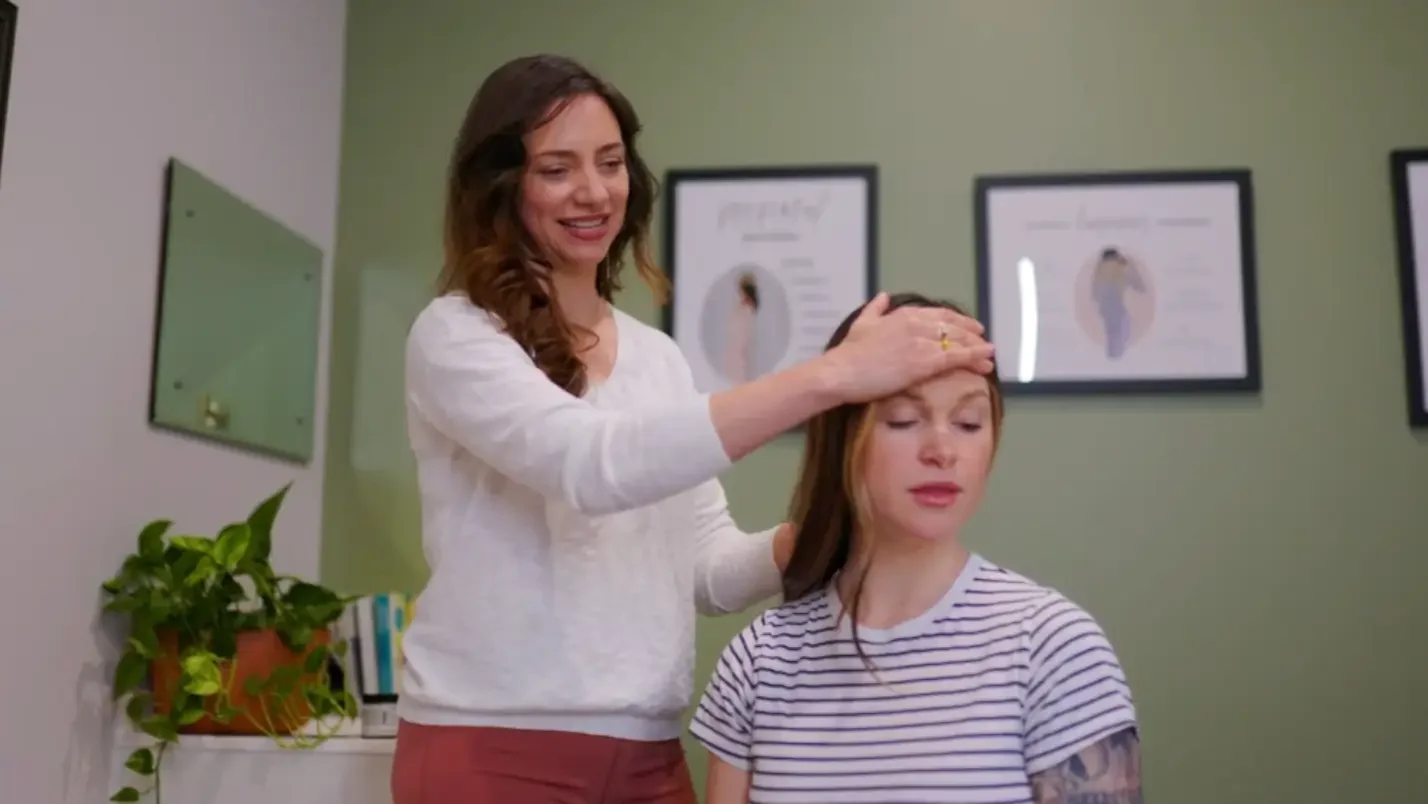 A woman performs a head massage on another woman seated in an office setting, with framed posters on the wall and a plant on a shelf.