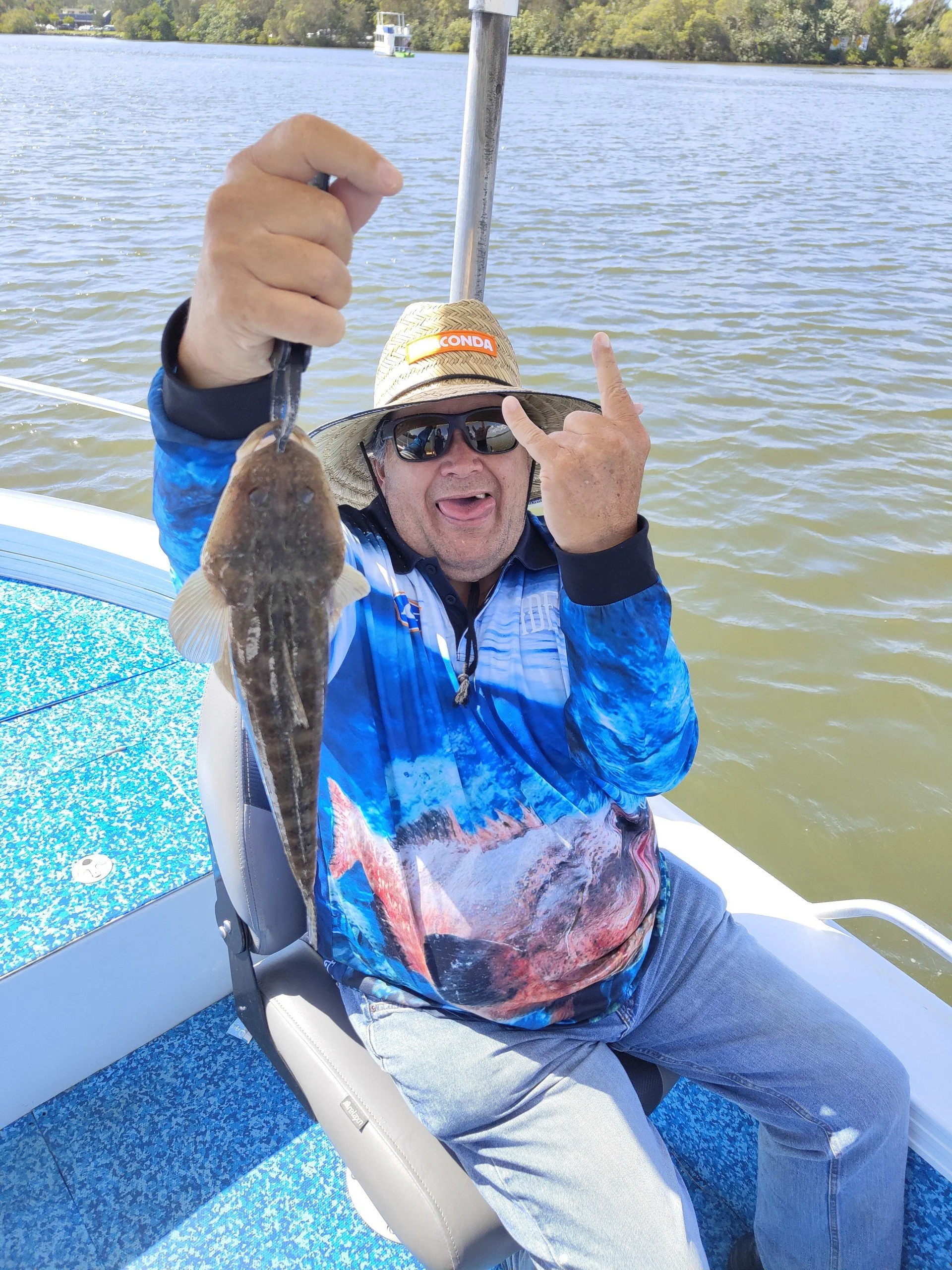 A man sitting on a boat holding a fish in his right hand and making a rock and roll hand gesture with his left hand. He is wearing sunglasses, a straw hat, and a colorful fishing shirt. The boat is on a body of water with trees in the background.