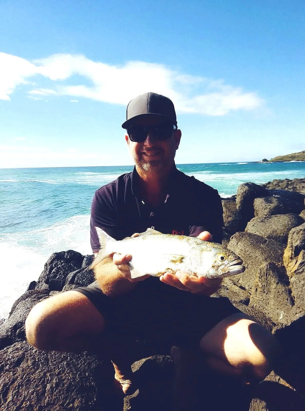 A man wearing a cap, sunglasses, and a dark shirt is sitting on rocks by the ocean, holding a fish he caught. The ocean waves are visible in the background, along with a clear blue sky and some clouds.
