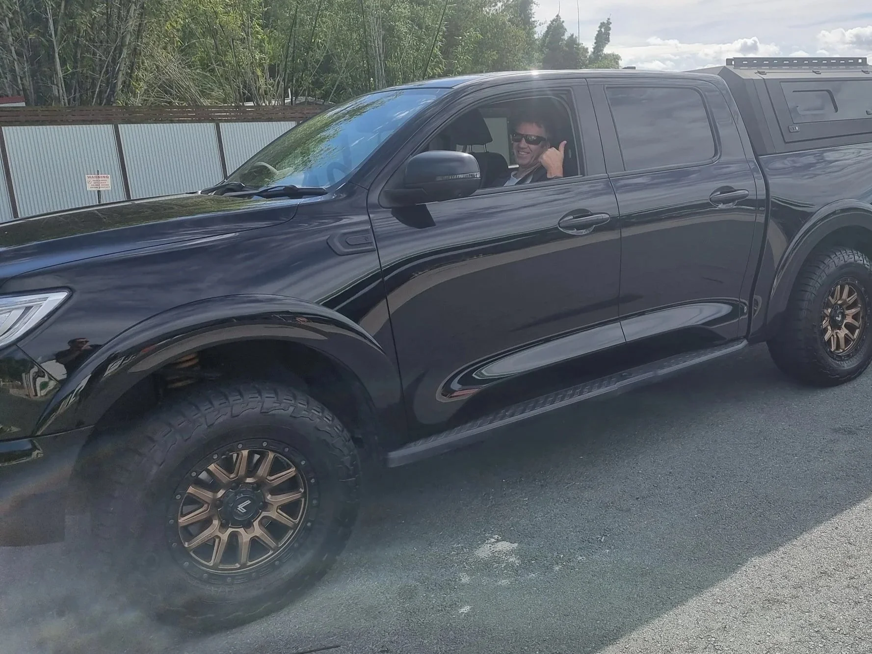 A black pickup truck with a camper shell parked on a gravel surface, towing a boat on a trailer. The driver, visible through the open window, is giving a thumbs-up and smiling.