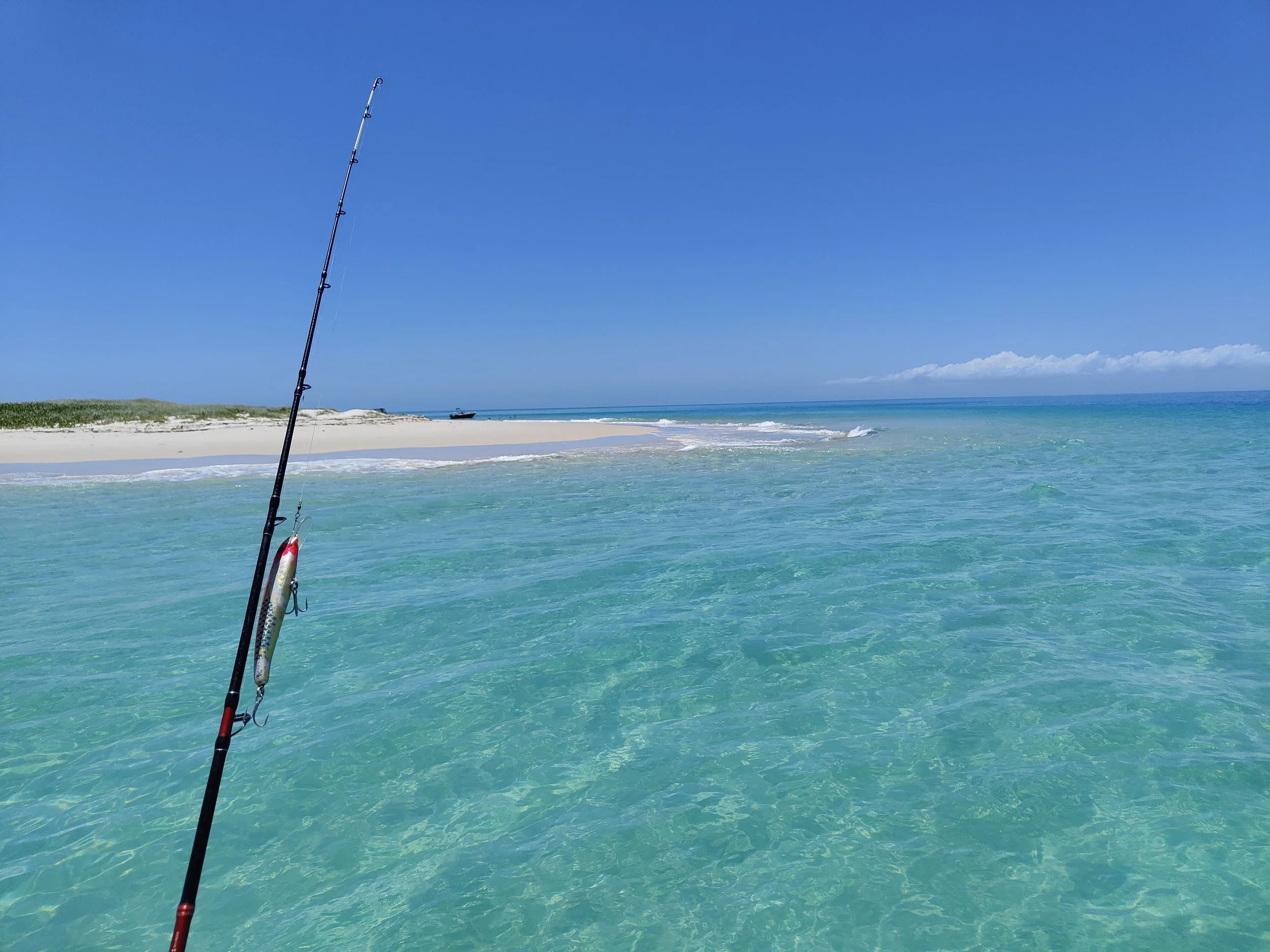 Fishing rod with a lure in clear turquoise water near a sandy beach under a blue sky.