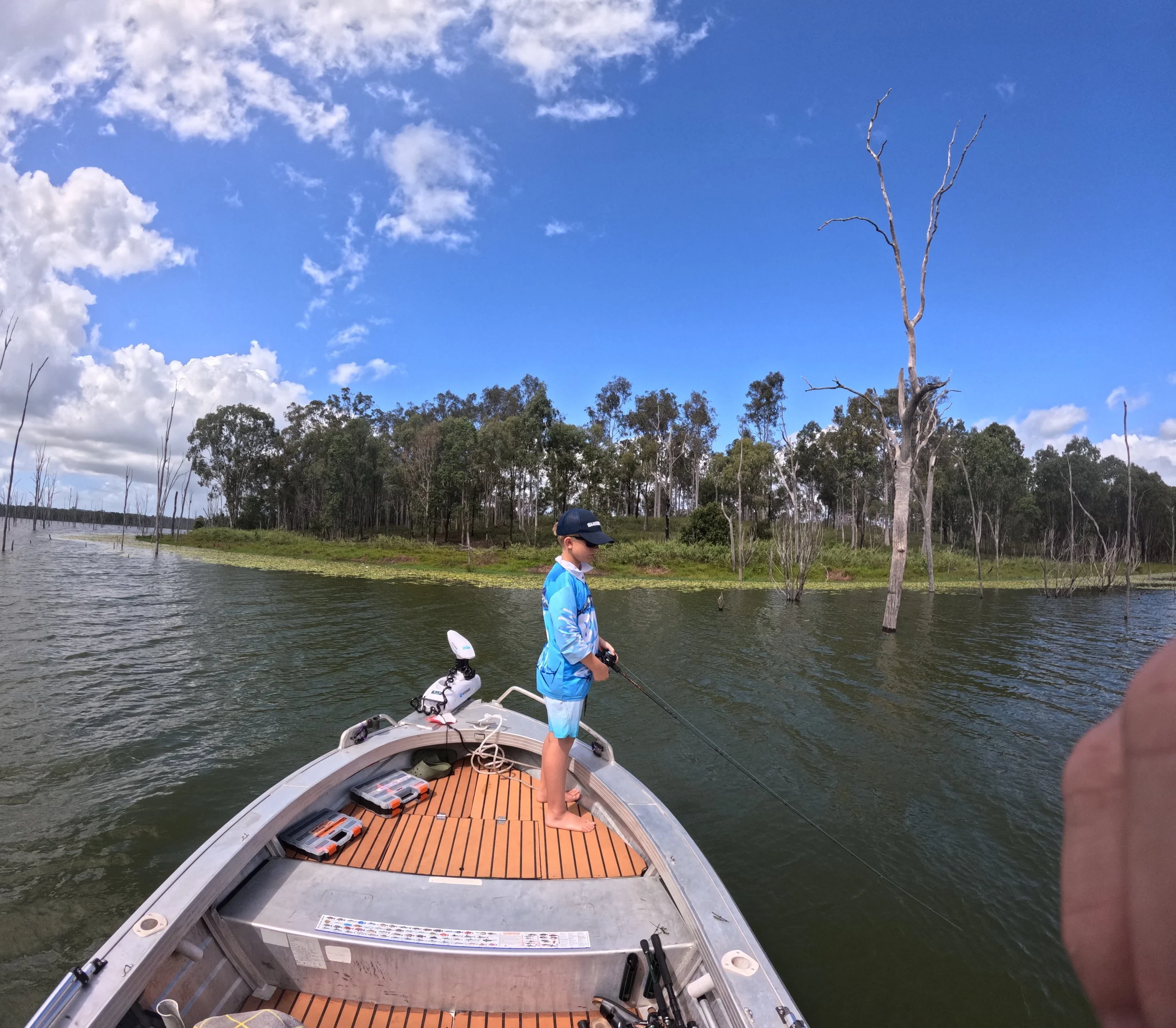 A boy in blue shirt and shorts standing on boat fishing in a lake with trees and blue sky in the background.