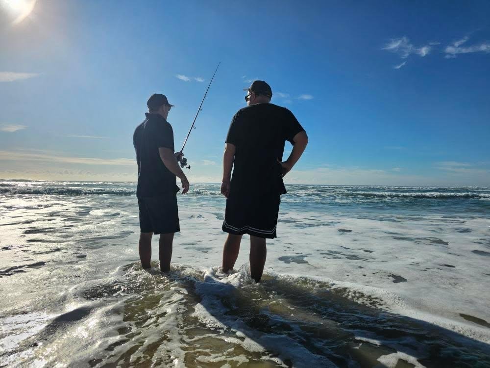 Two men standing in the ocean waves on a beach, with one holding a fishing rod, under a partly cloudy blue sky.