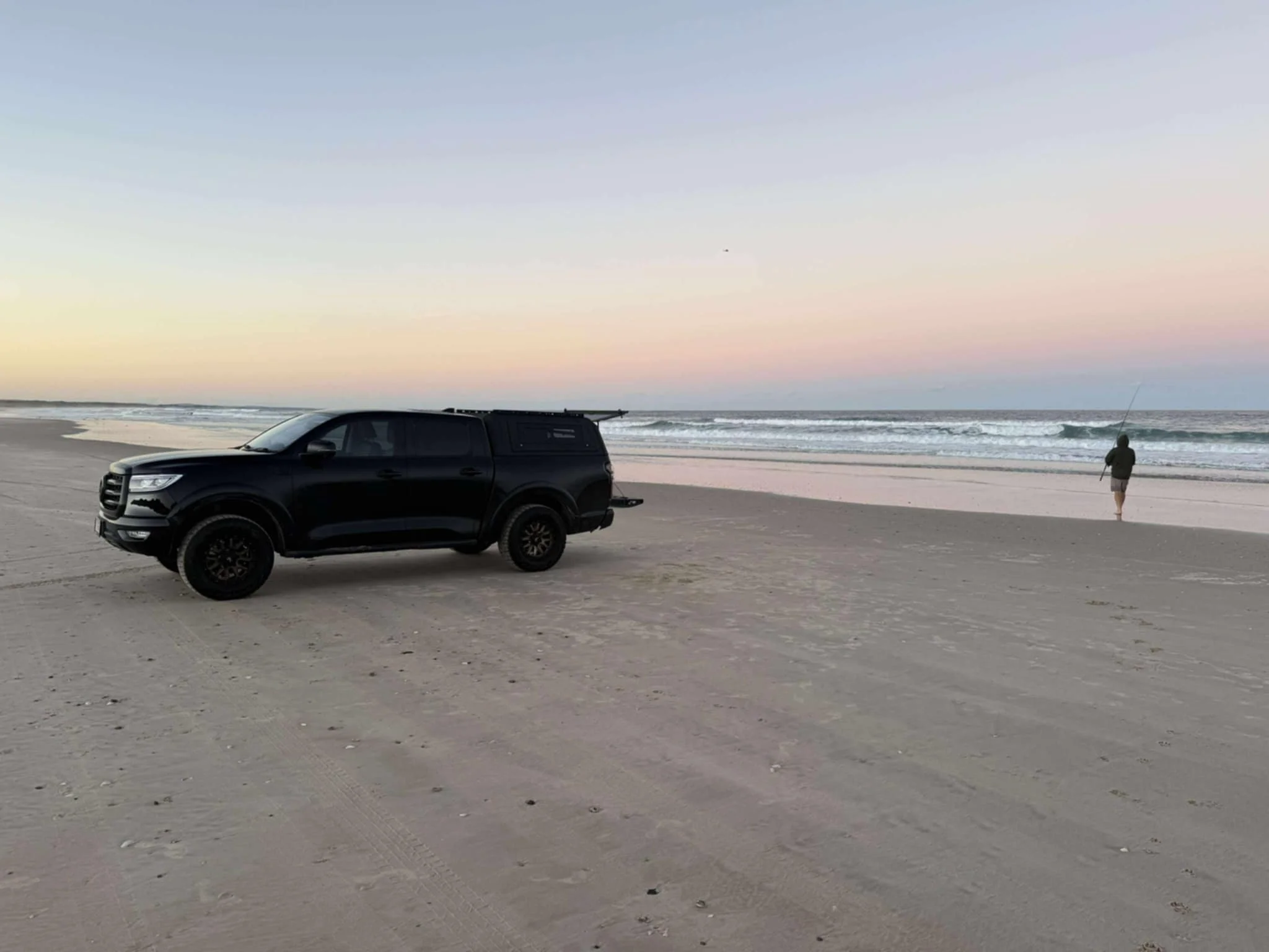 A black pickup truck parked on the sandy beach with a person fishing near the shoreline at sunset.